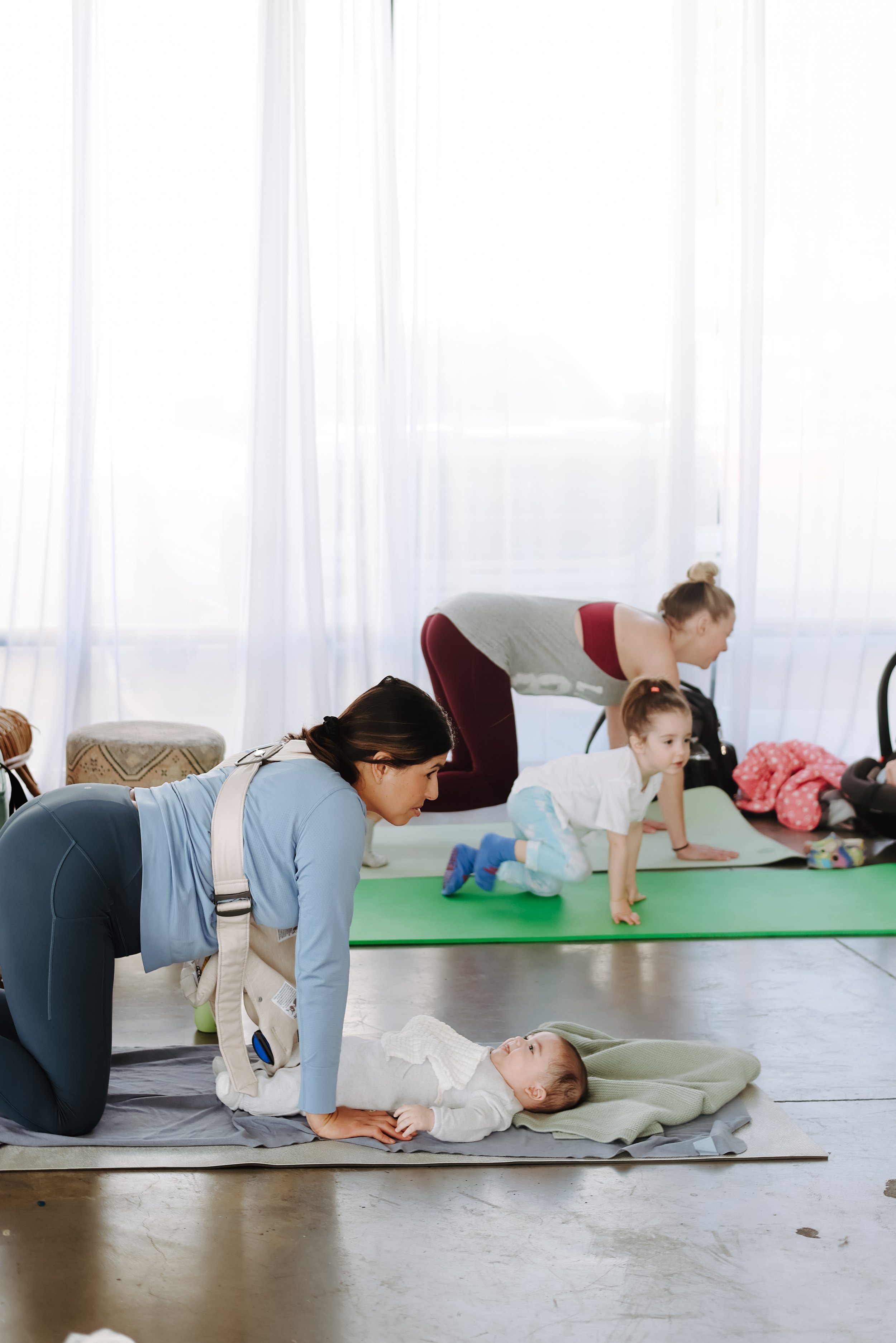 Infant participating in a prenatal yoga class with instructor, other children and adults practicing yoga in bright room with sheer curtains.