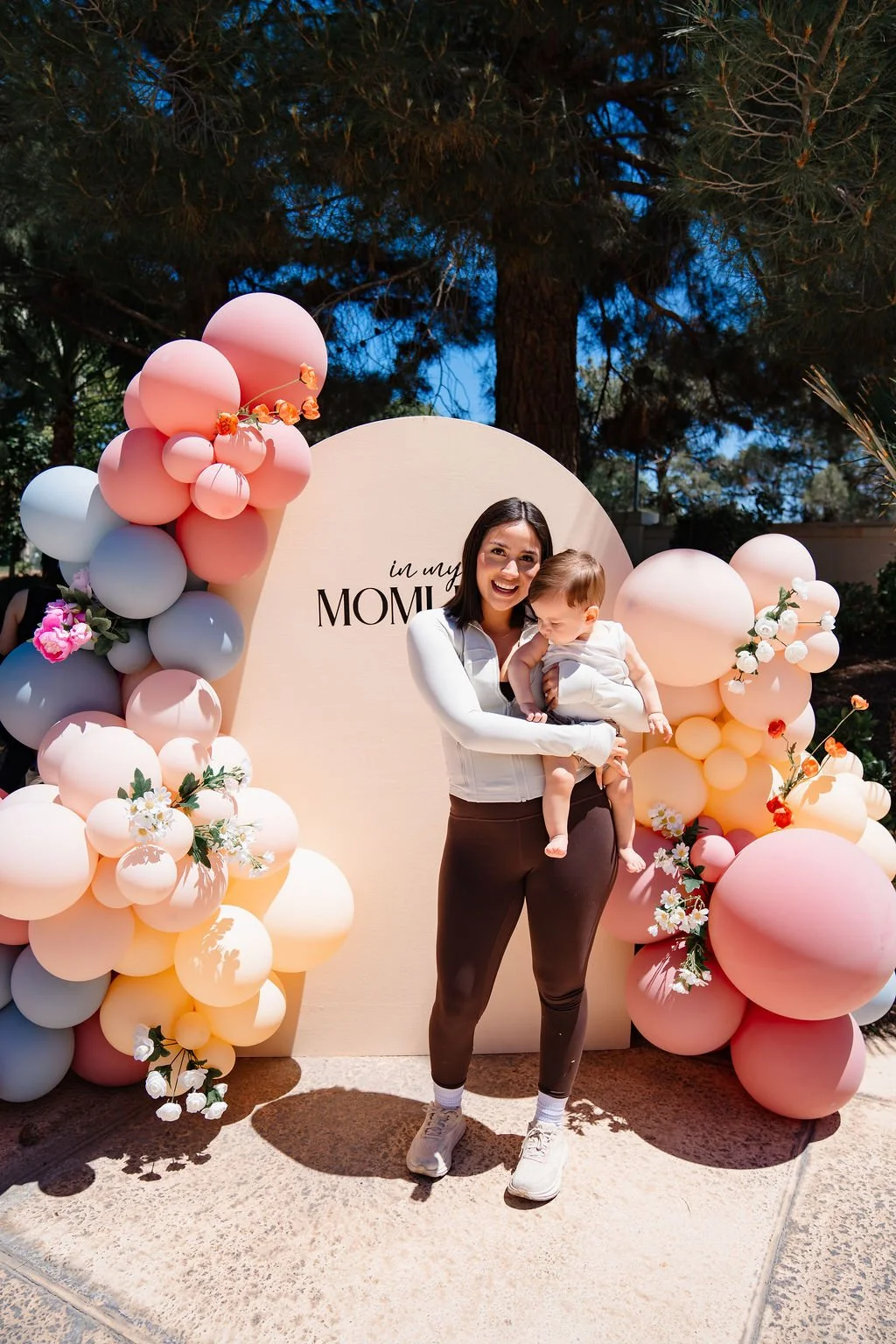 A smiling woman holding a toddler in front of a pastel-colored balloon display with flowers, at an outdoor event with trees and blue sky in the background.