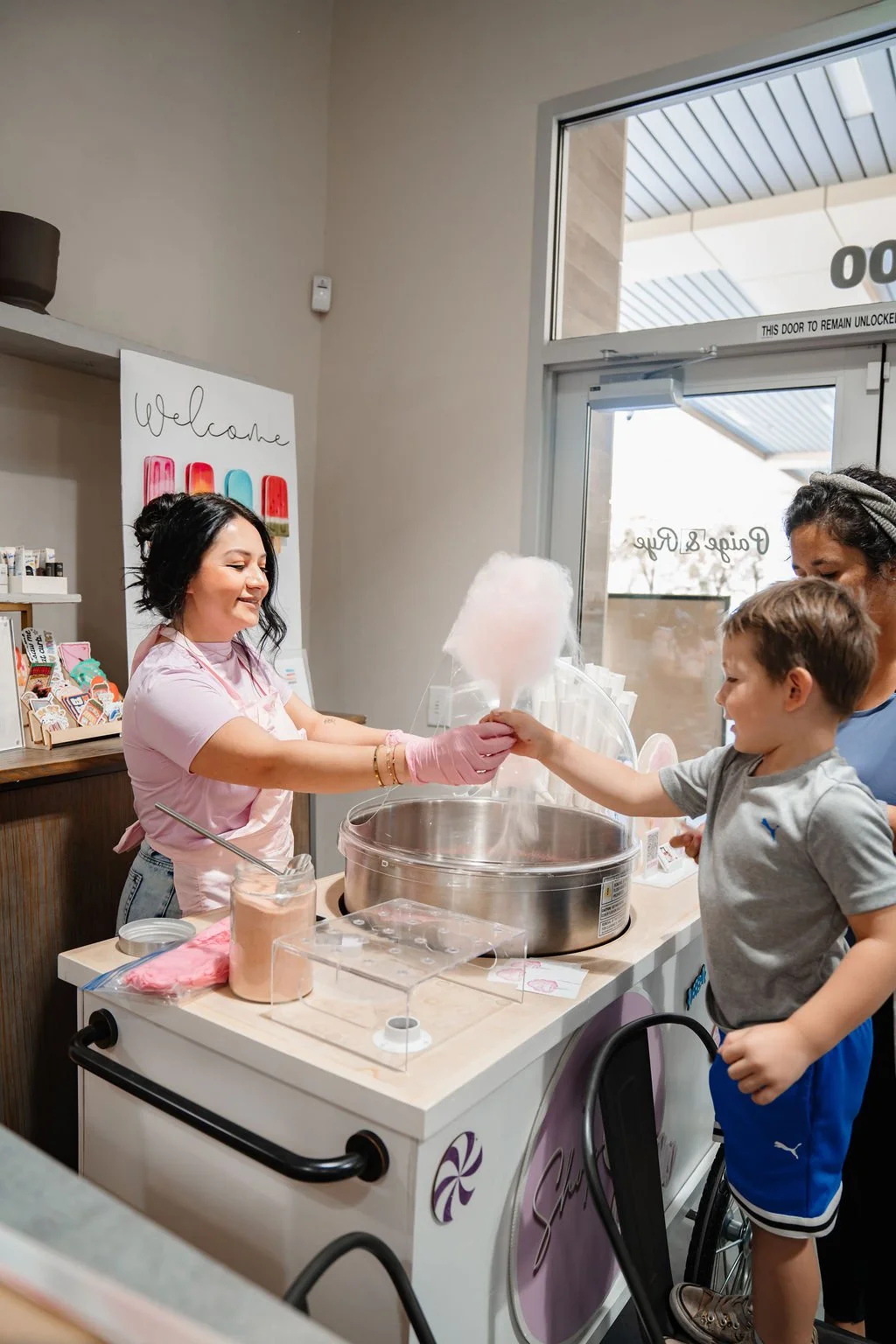 A woman serving cotton candy to a young boy at a cotton candy stand, with another woman nearby, inside a shop with a 'Welcome' sign and a glass door in the background.