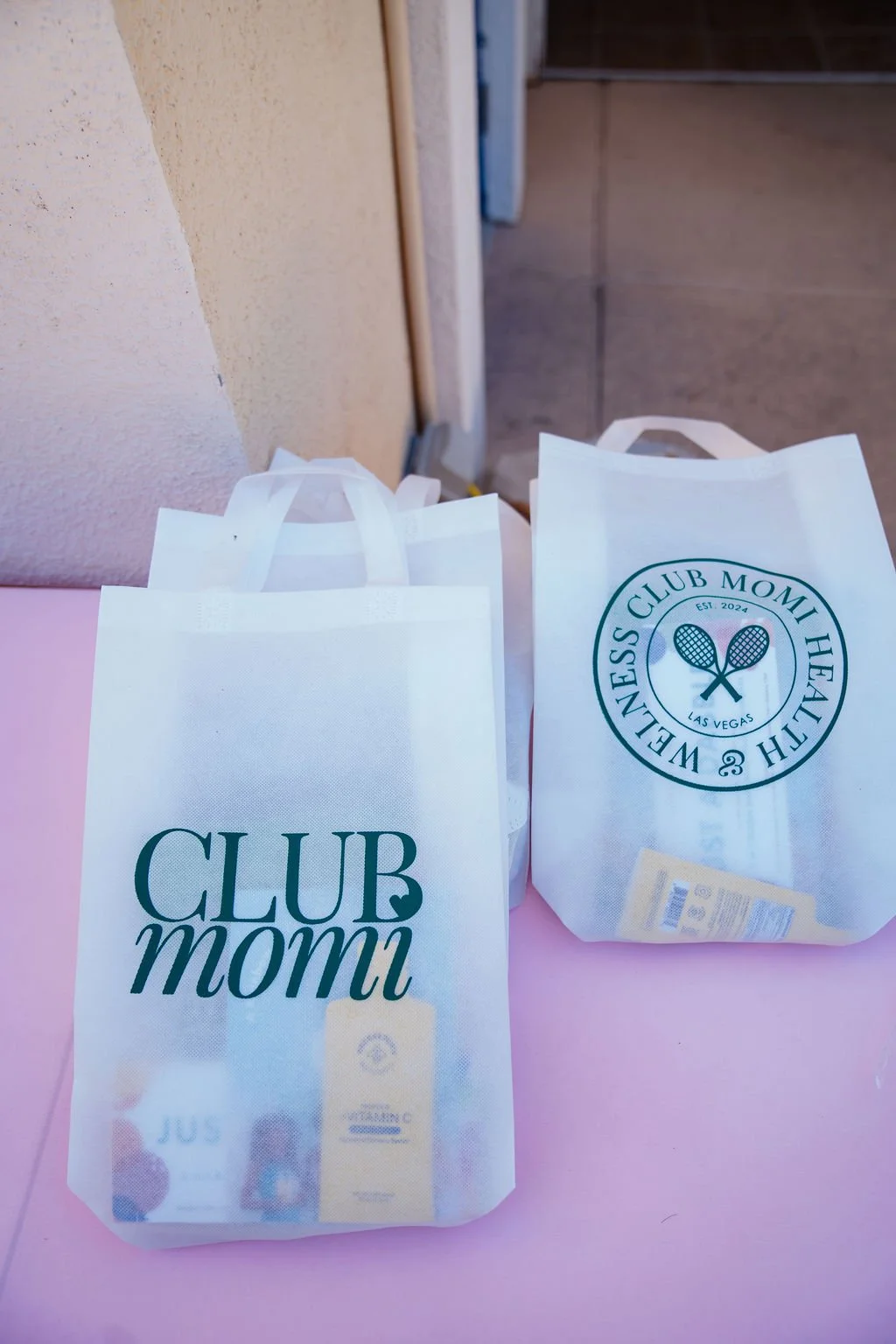 Two white paper bags with 'CLUB momi' logo, placed on pink surface near beige wall and doorway.