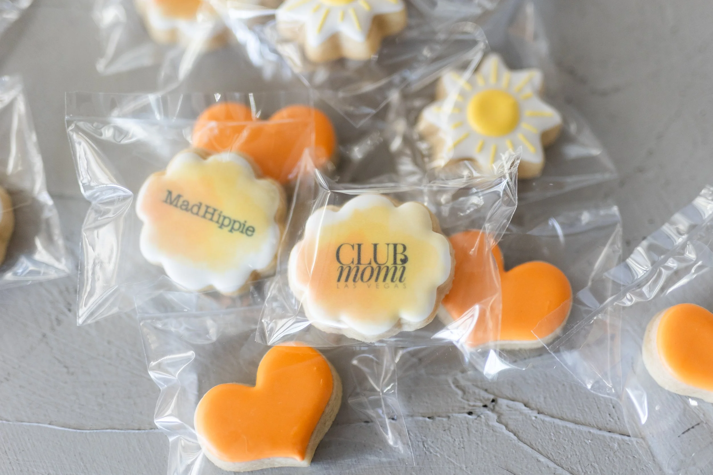 Assorted decorative cookies in clear packaging, shaped like hearts, flowers, and suns, with some displaying text reading 'Mad Hippie' and 'CLUB mom! LAS VEGAS.'