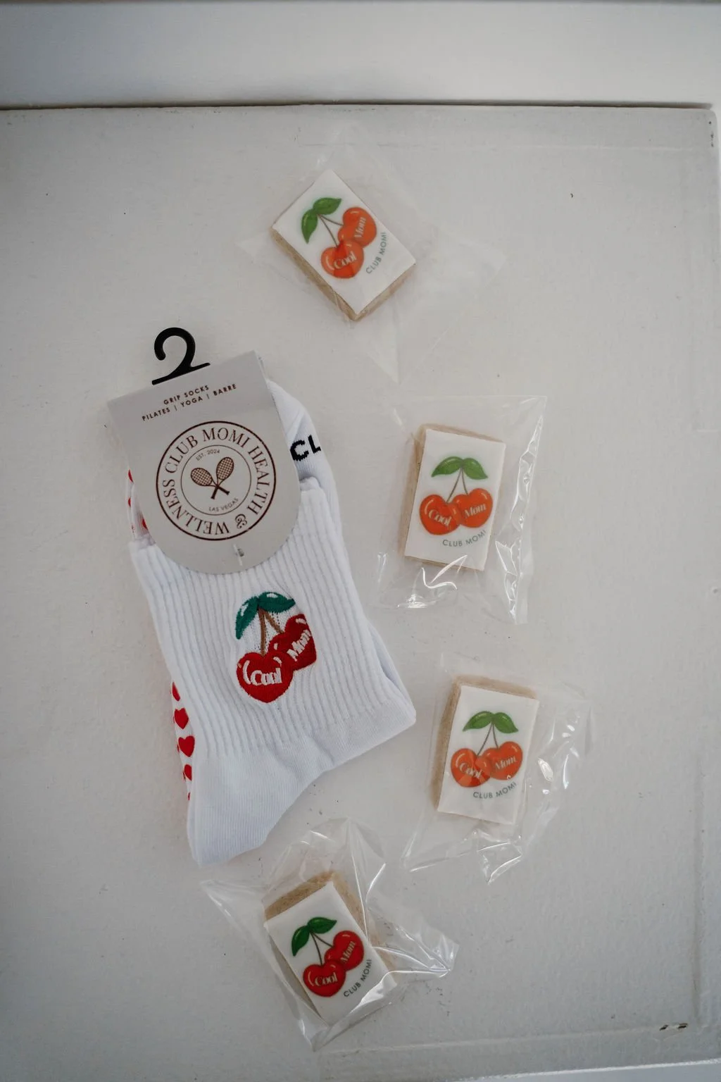 A white sock with cherry graphics and teeth, and multiple small white erasers with cherry logos, all on a white surface.