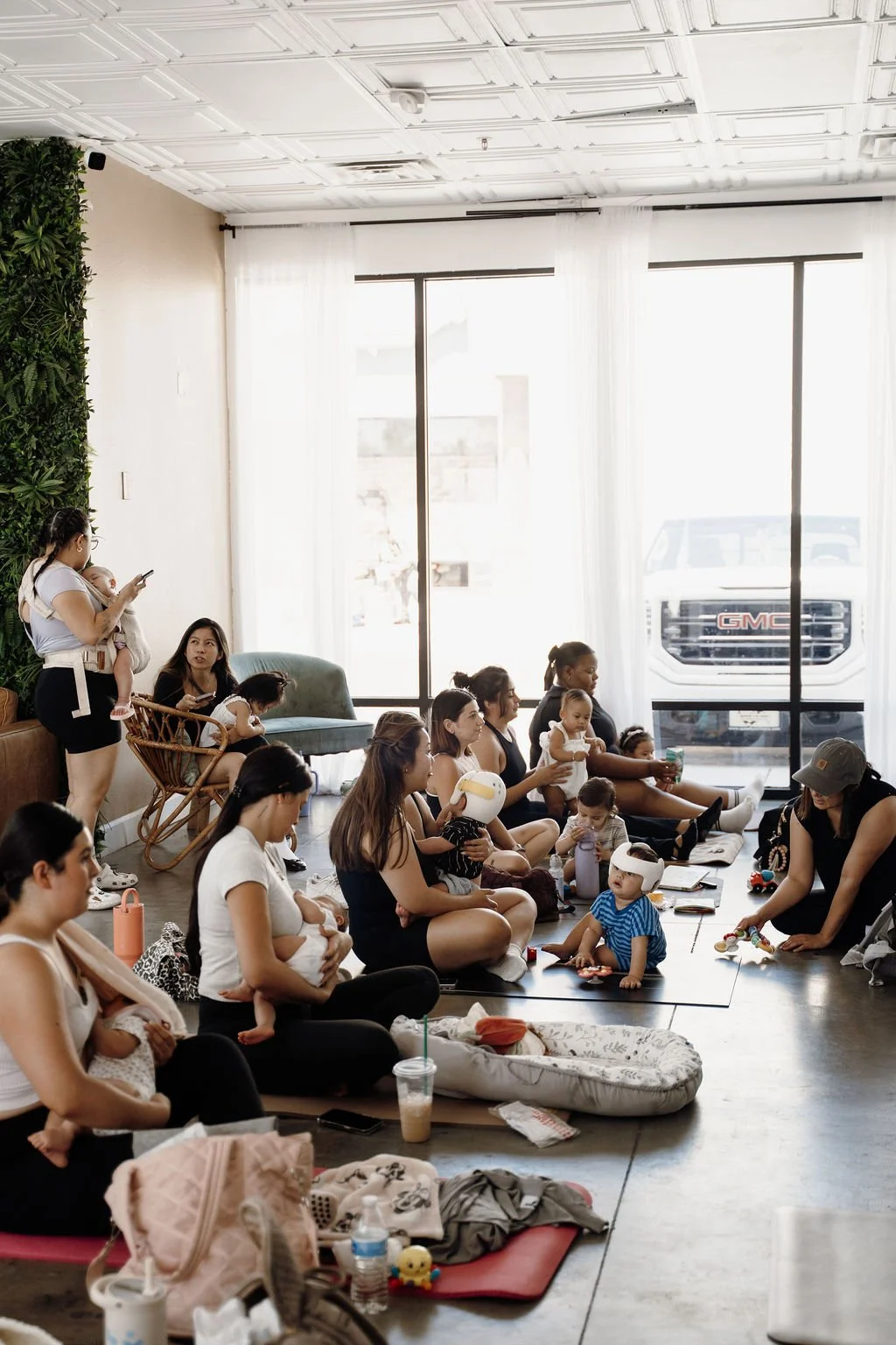 Group of women and children sitting on the floor and chairs during a daytime indoor gathering in a bright room with large windows, some wearing virtual reality headsets, with a mostly empty space in the foreground.