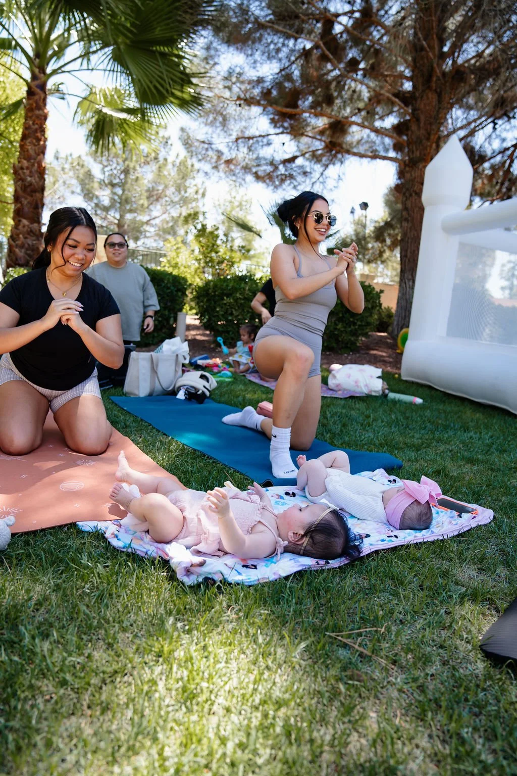 Women and children enjoying a sunny outdoor gathering, with two women kneeling and a woman doing a yoga pose on a mat while kids lie on blankets on the grass.