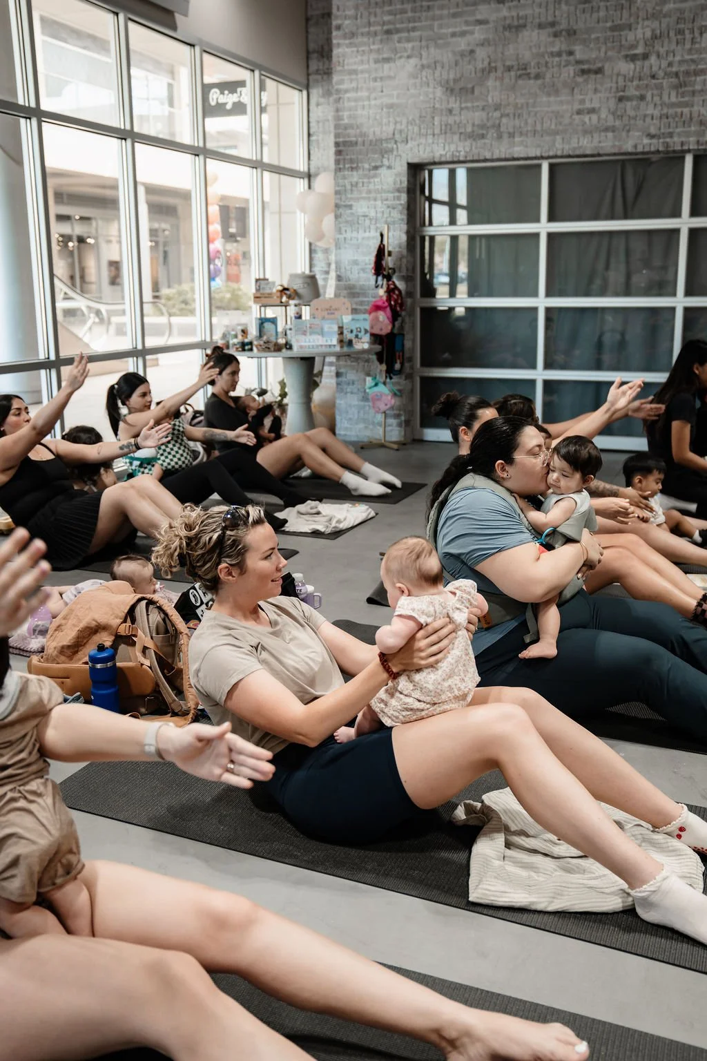 Group of women and children participating in a seated activity or class inside a modern space with large windows and exposed brick walls.