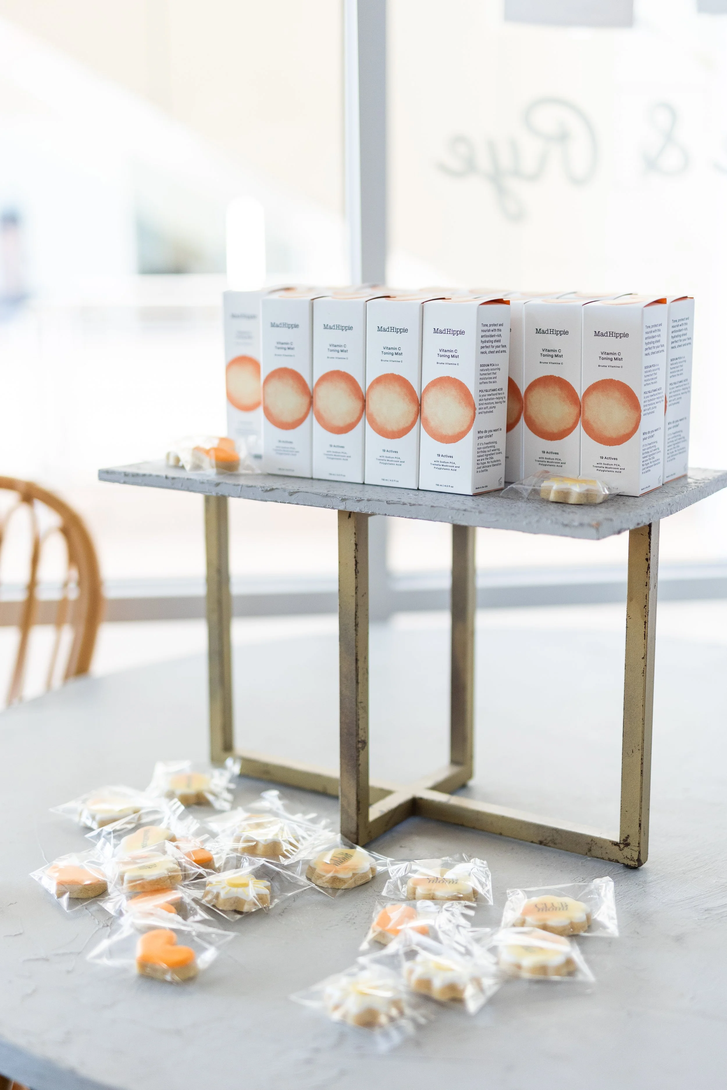 Display of boxes of Mad Hippie Vitamin C Toning Mist on a table with individually wrapped cookies in front.