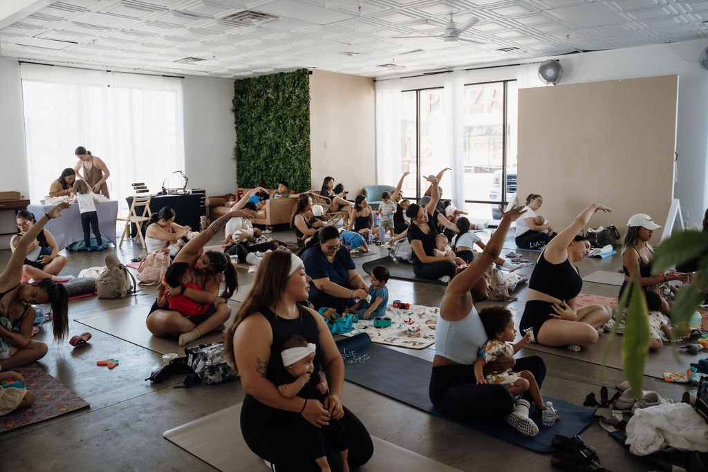 Group of women and children practicing yoga in a bright, spacious studio with large windows, a green wall decoration, and calming indoor plants.