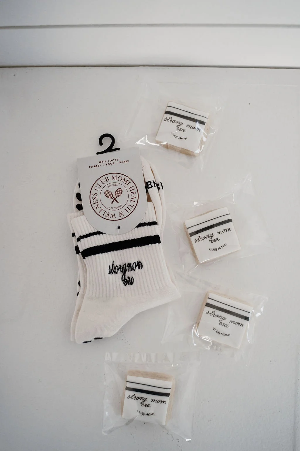 A collection of white socks with black stripes and handwritten text, individually packaged in plastic, along with a pair of grip socks labeled 'Strong Mom Era,' all displayed on a white surface.