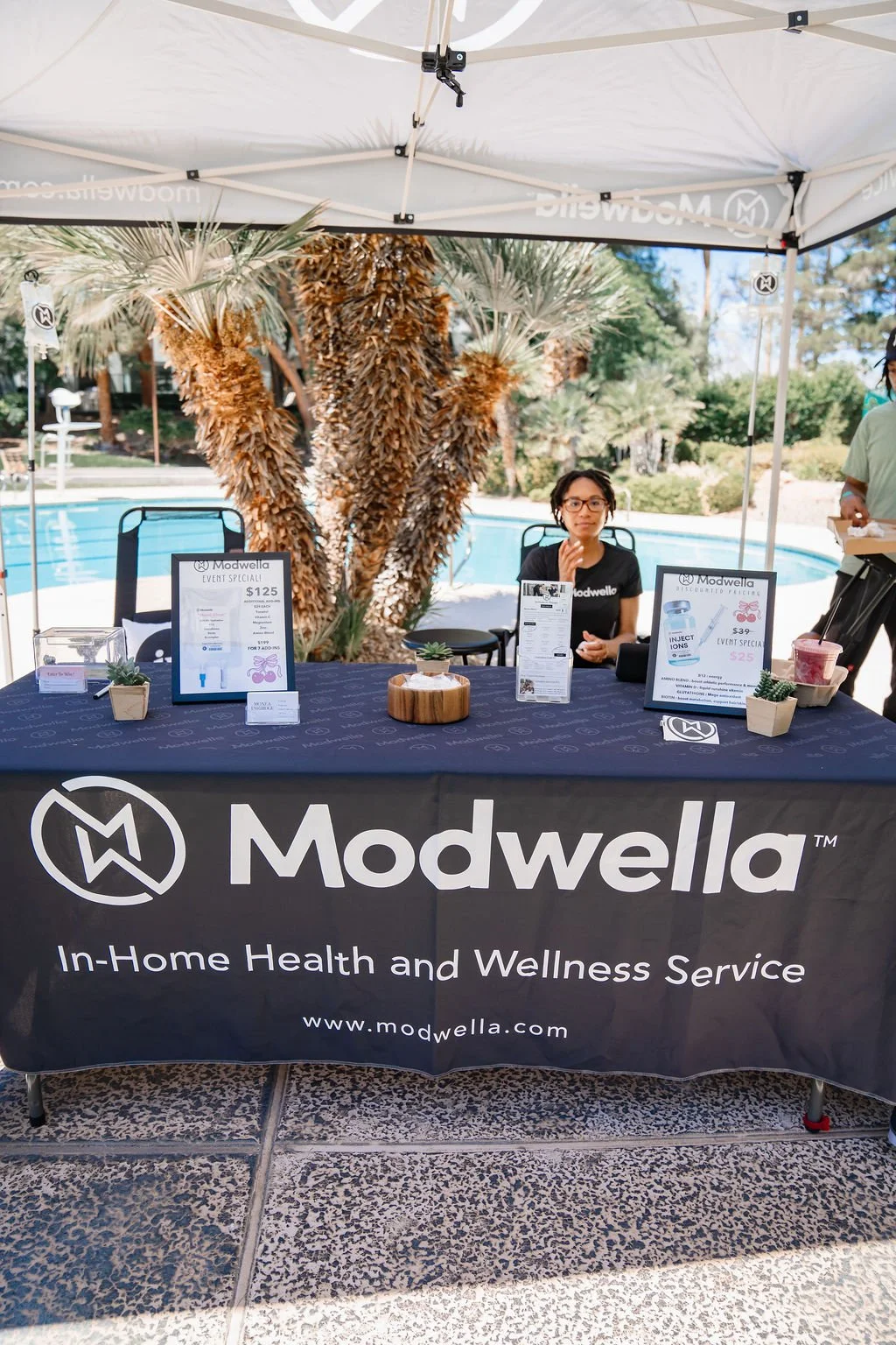 A woman sitting at a booth for Modwella, a health and wellness service company, with a pool and palm trees in the background.