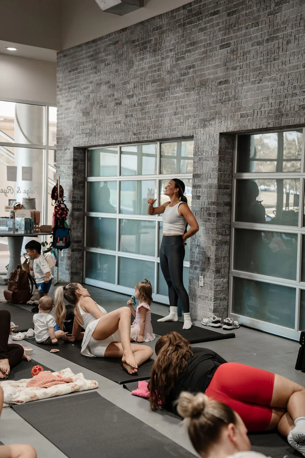 A woman leading a fitness class with children and adults doing exercises on mats in a gym.