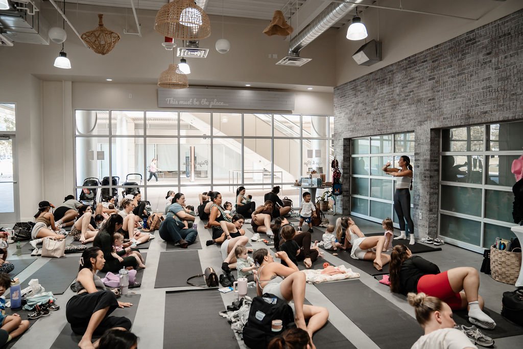 A large group of women and children participating in a fitness or yoga class indoors, with a woman leading at the front and others on yoga mats.