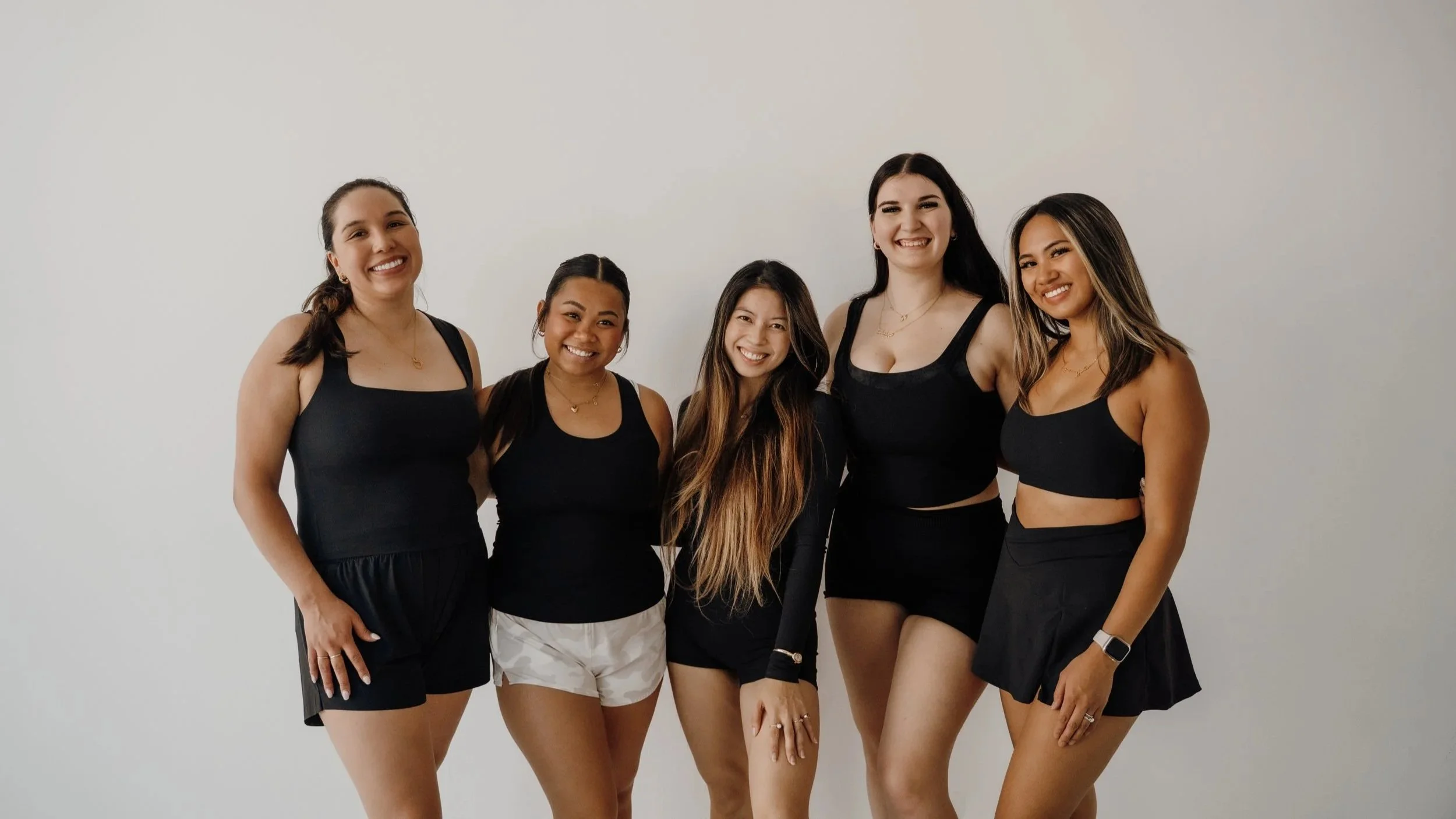 Five women standing together smiling against a plain white wall, wearing black athletic wear.