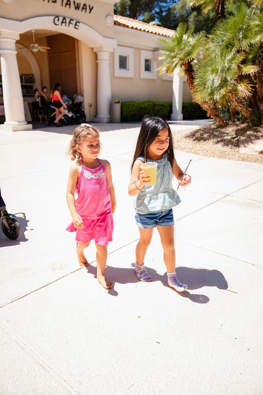 Two young girls walking outdoors in front of a building labeled 'Eataway Cafe'. One girl, wearing a pink dress, is barefoot, and the other, in a gray top and denim shorts, is holding a yellow drink and a straw. They are smiling and enjoying the sunny