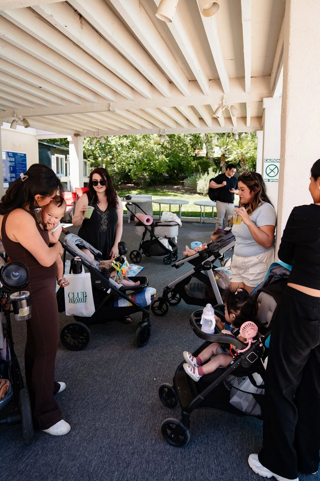Group of women and children gathered under a shaded outdoor area, some sitting in strollers, others standing and talking, with trees and greenery in the background.