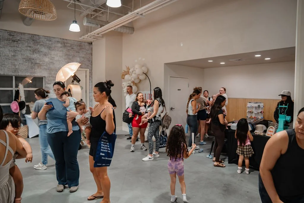 A group of women and children gathered in a modern indoor space with high ceilings, bright lighting, and minimal decor, some standing and chatting, others near a table with items on display.