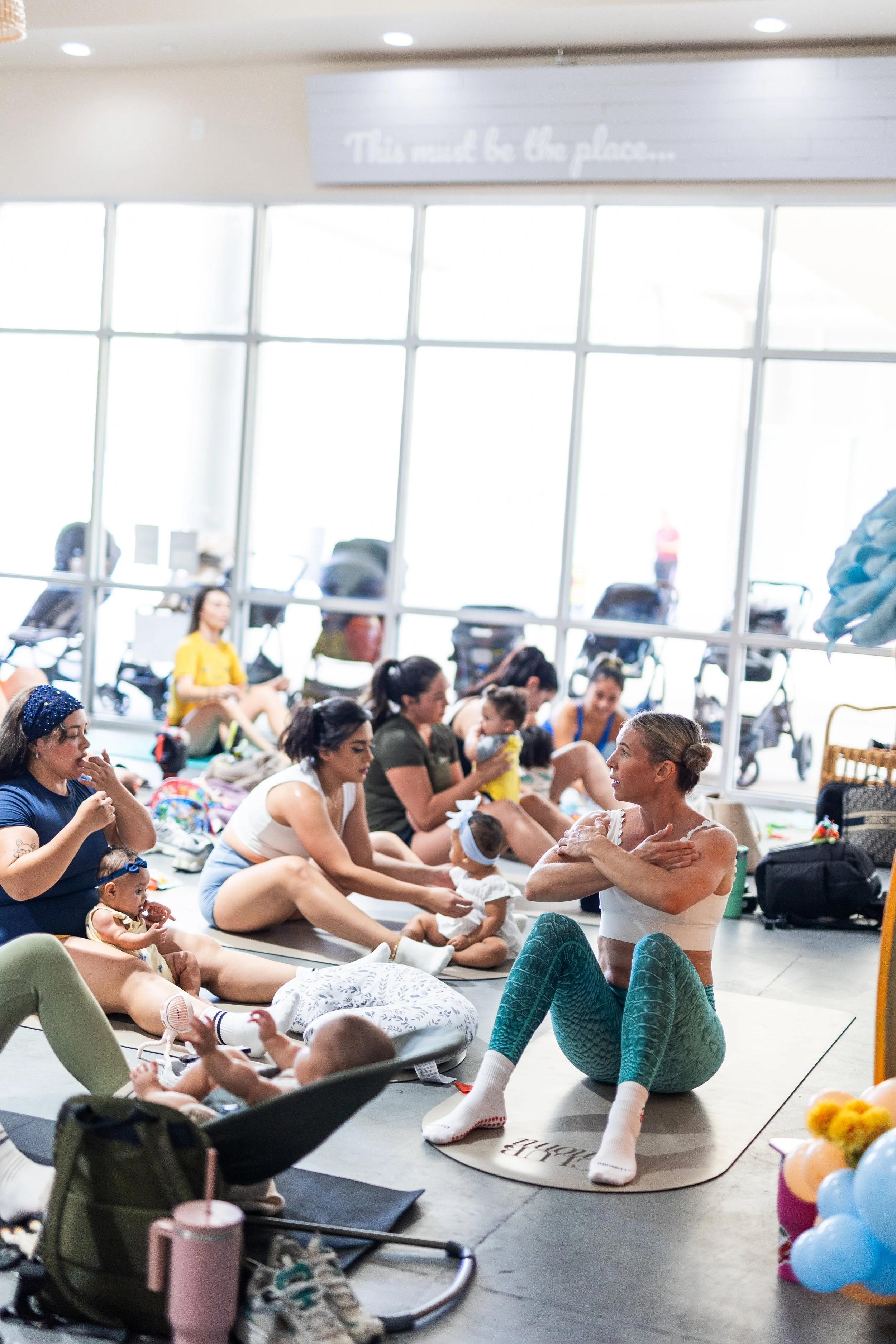 A group of women and babies participating in a prenatal exercise class in a bright room with large windows. A woman is demonstrating stretches while others, some with babies, follow along.