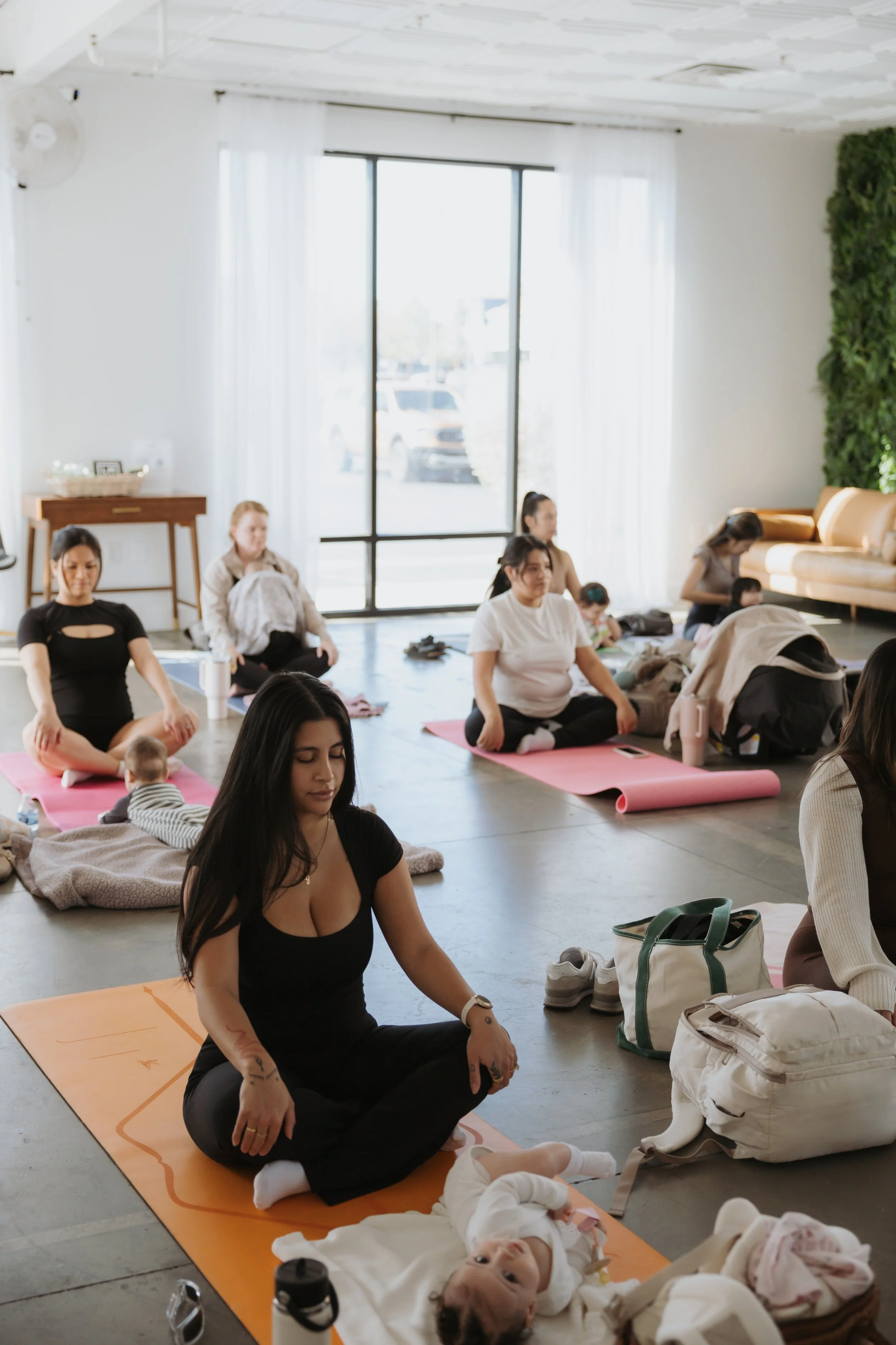 A group of women and babies participating in a yoga class in a bright, airy studio with large windows and white curtains. The women are seated on yoga mats, some with infants nearby.
