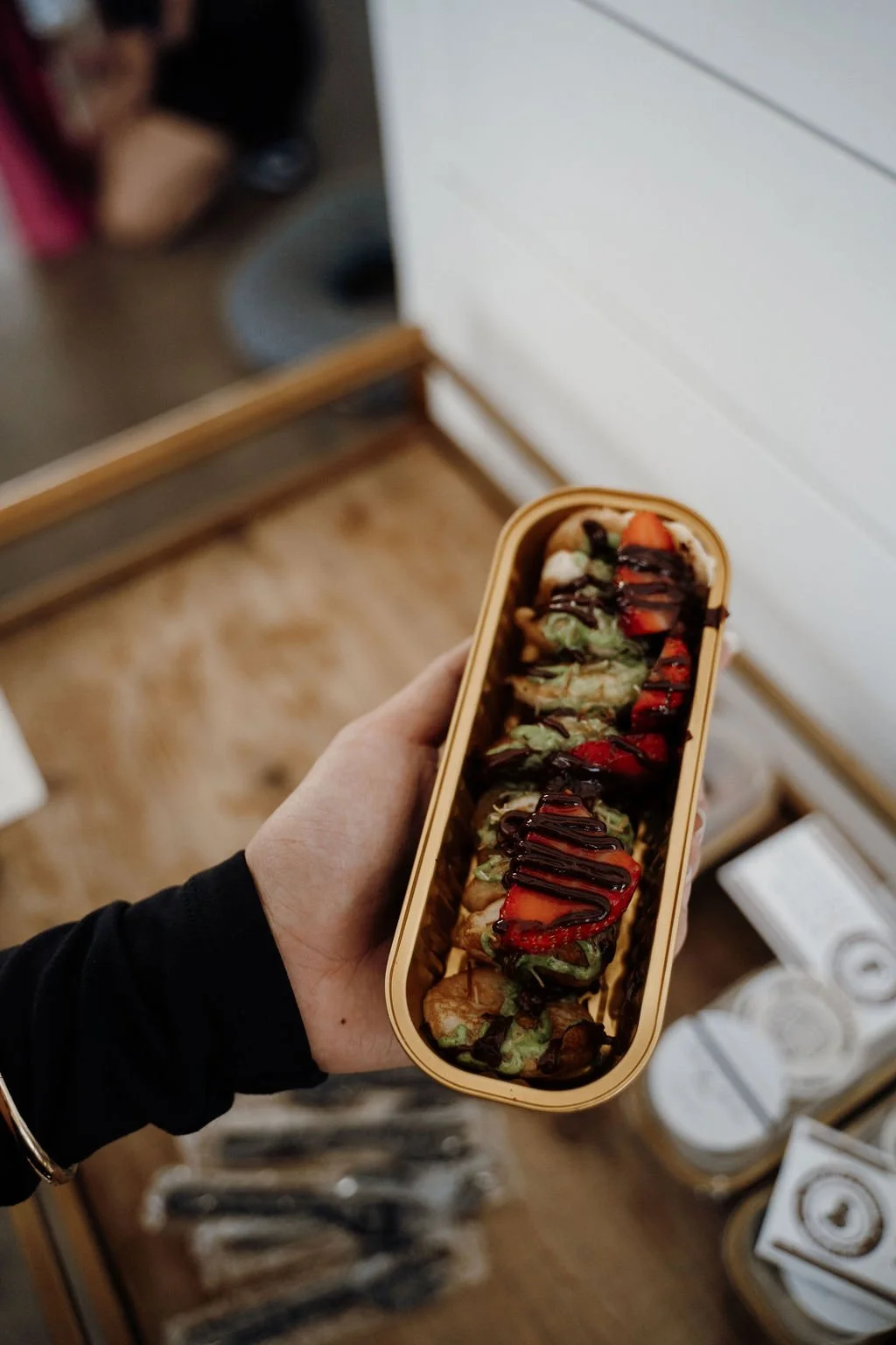 A hand holding a rectangular container of sushi rolls drizzled with chocolate and topped with strawberries.