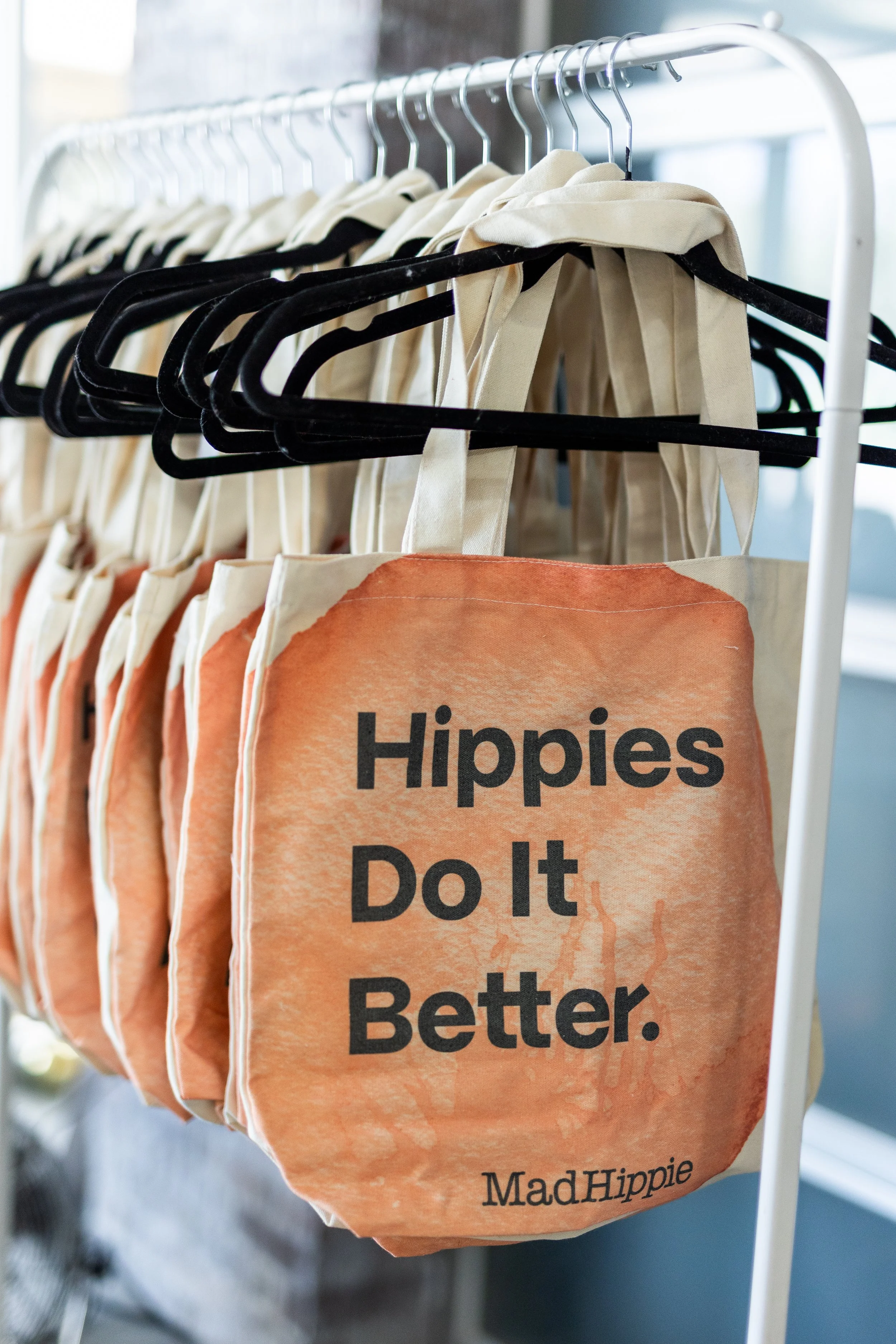 A row of beige tote bags hanging on a white rack, with one tote bag in front displaying the text 'Hippies Do It Better.' and 'Mad Hippie' printed at the bottom.
