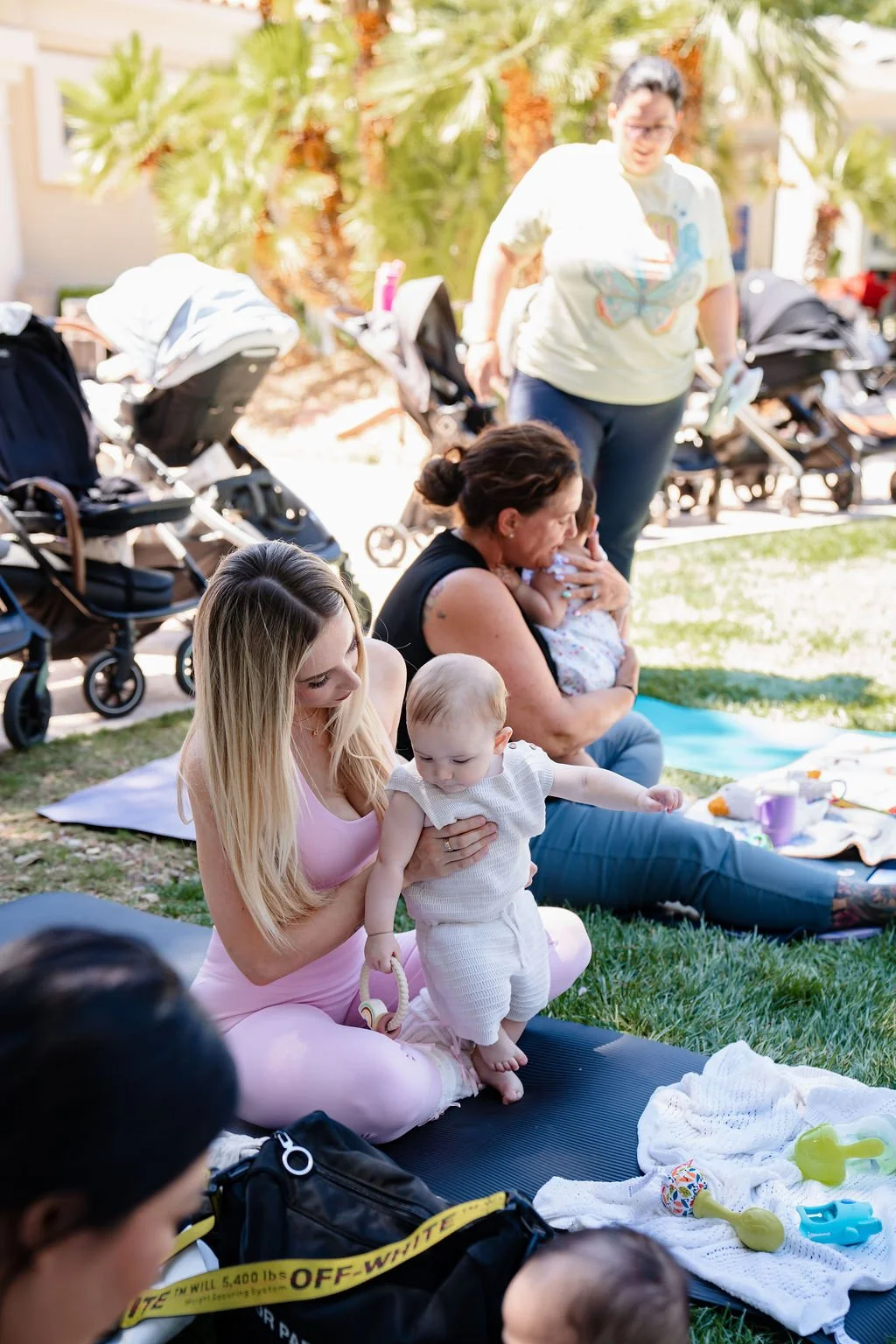 Group of women with babies sitting on a grassy area during a gathering or outdoor event, with strollers and palm trees in the background.