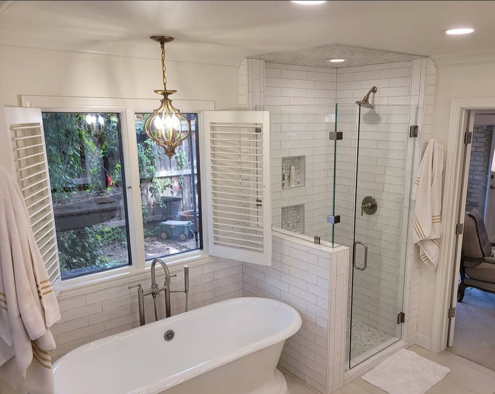 A modern bathroom featuring a standalone bathtub beneath a window with plantation shutters, a glass-enclosed shower with white subway tile, and a decorative chandelier hanging from the ceiling.