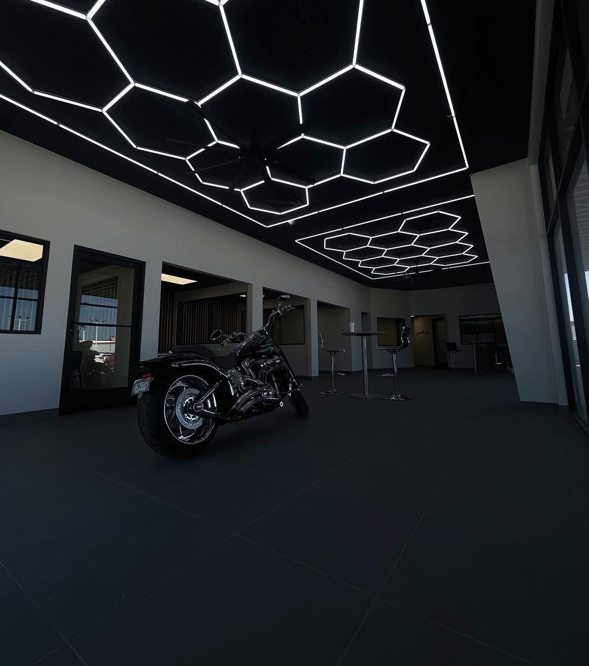 A black motorcycle inside a modern room with white walls and illuminated hexagonal and honeycomb pattern ceiling lights.
