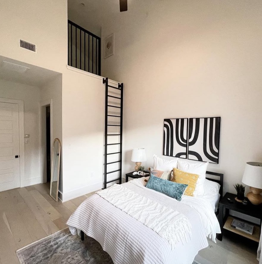 Bedroom with a bed, pillows, and nightstands with lamps, a black ladder leading to a loft area, and artwork hung on the wall above the bed.
