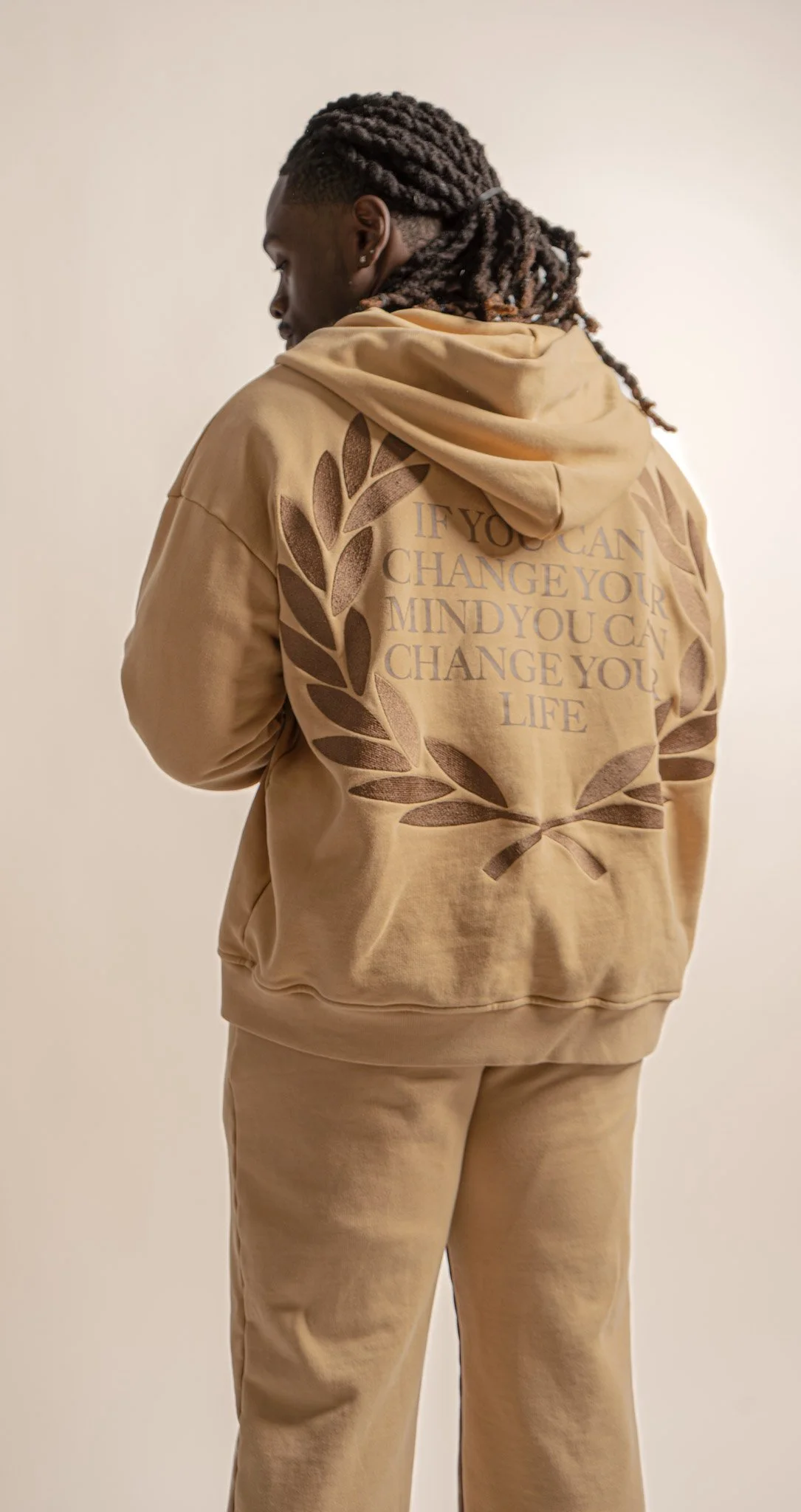 Back view of a person wearing a tan hoodie with a quote and laurel wreath design, standing against a plain background.