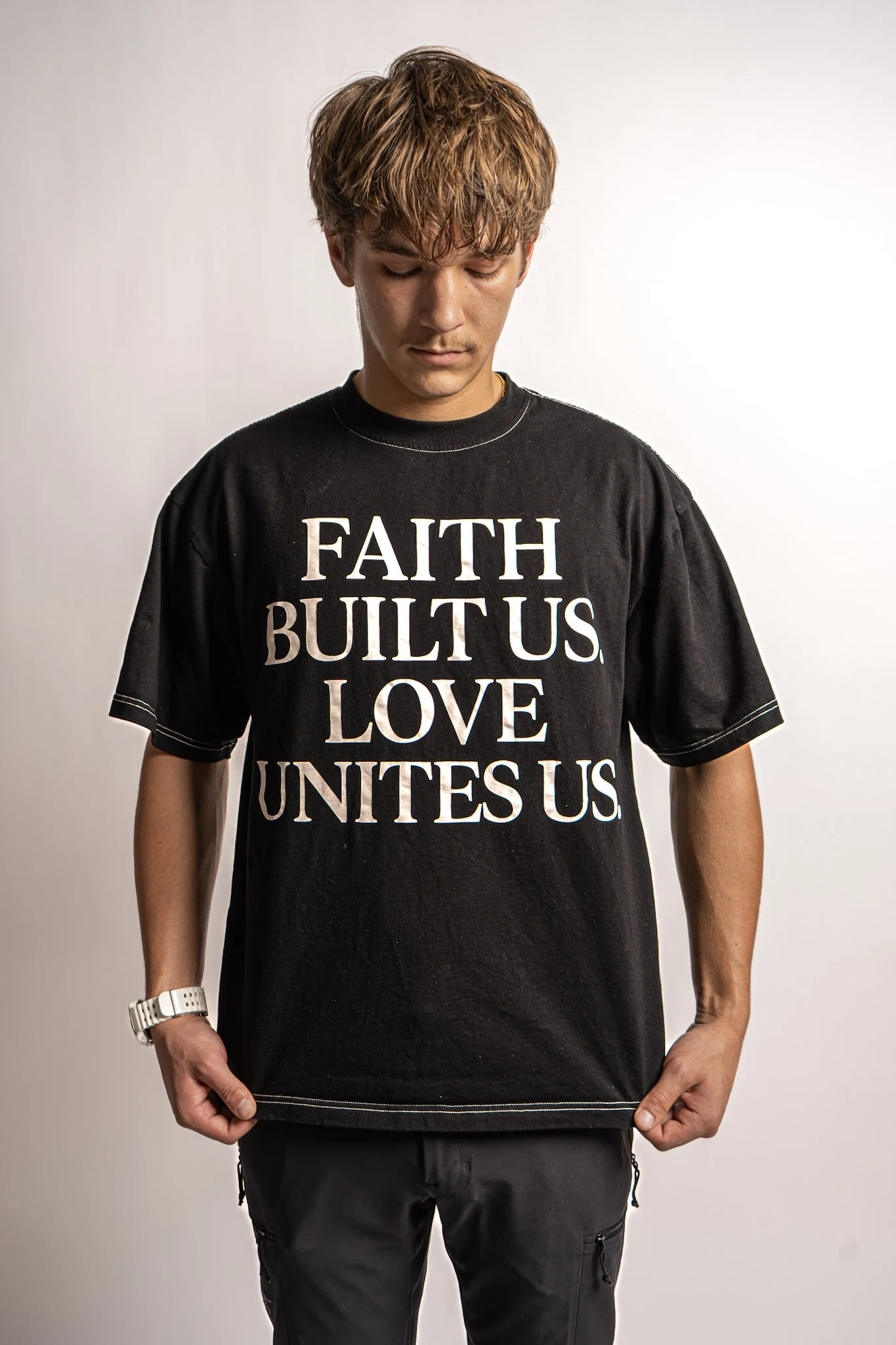 Young man with brown hair looking down, wearing a black T-shirt with white text that reads 'FAITH BUILT US LOVE UNITES US'.