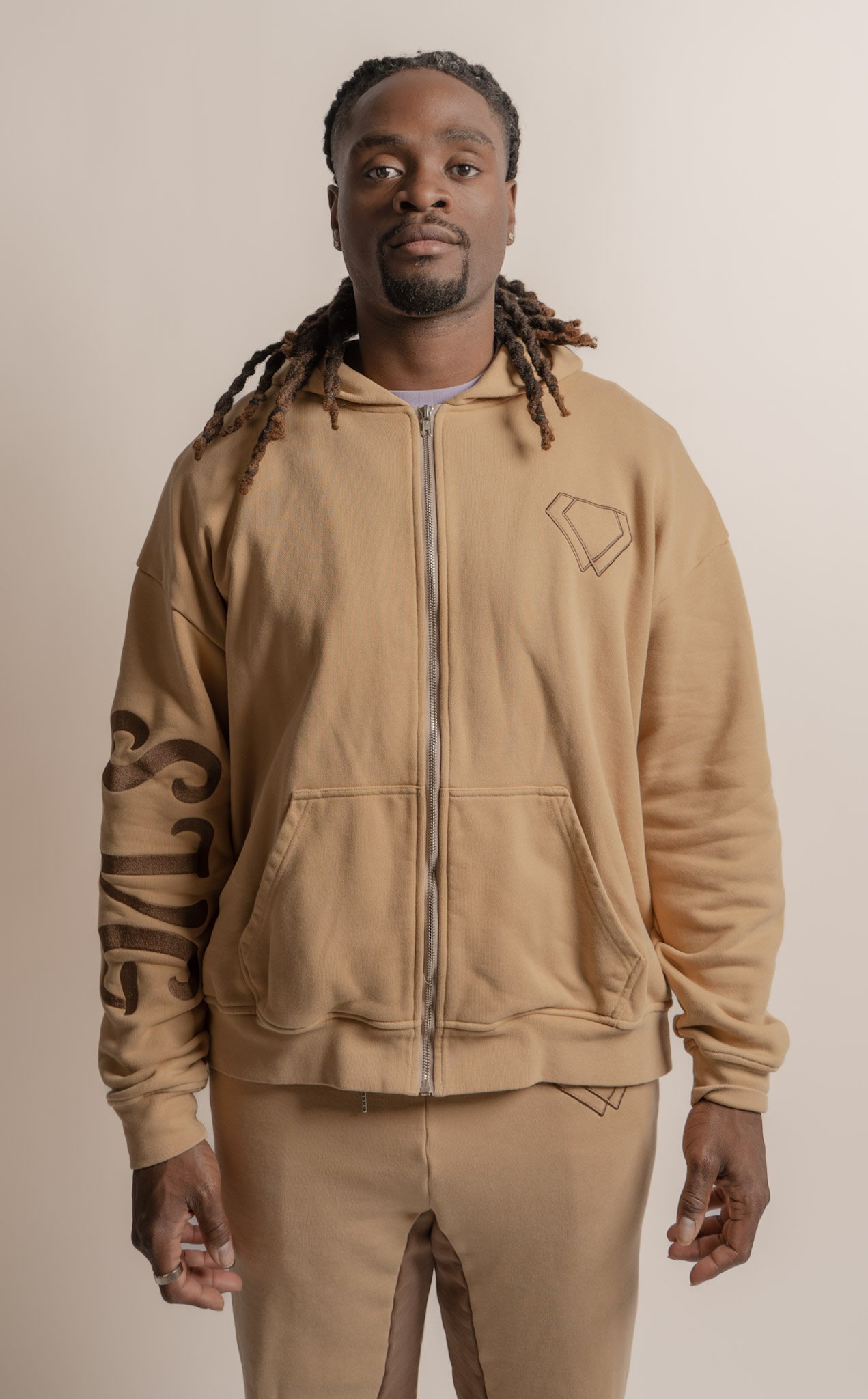 A man with long dreadlocks wearing a beige hoodie and matching sweatpants, standing against a plain light-colored background.
