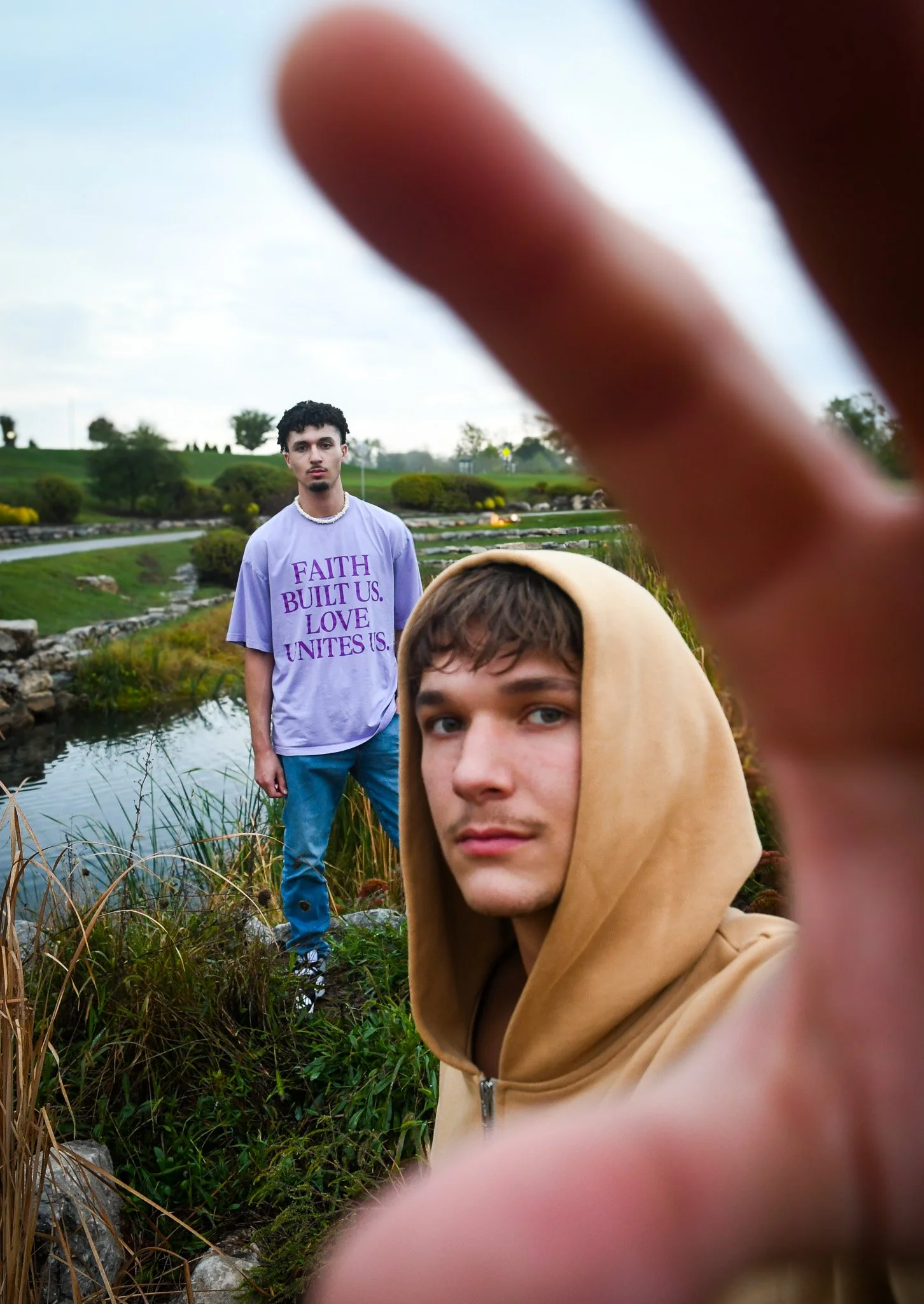 Two young men outdoors near a pond, one in the foreground wearing a beige hoodie looking at the camera, and one in the background wearing a purple T-shirt with text, standing near the water, with a hand partially blocking the view.