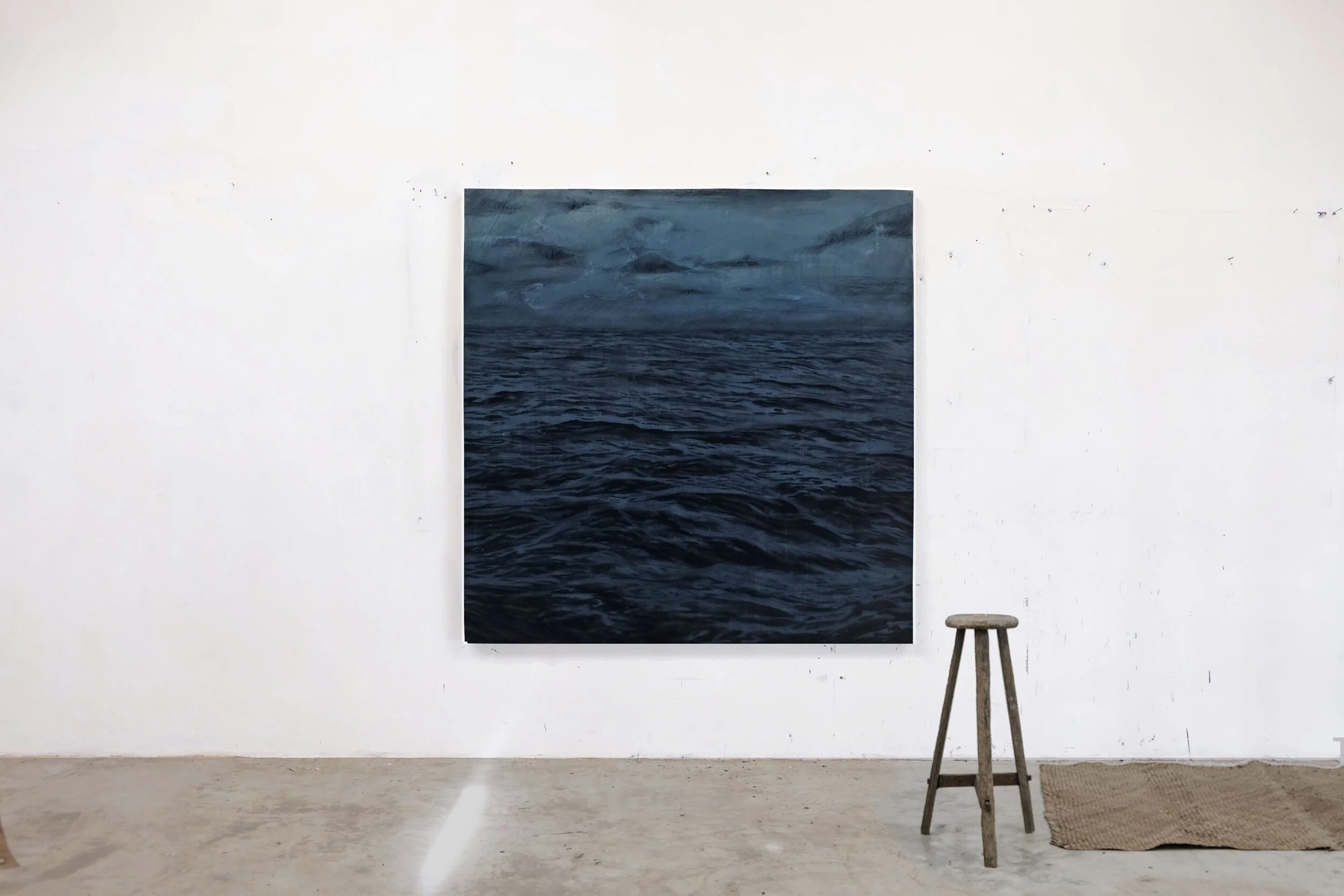 Cuadro con imagen de un mar con olas y cielo nublado, colgado en una pared blanca en una galería, con un taburete y un tapete de yute en el piso.