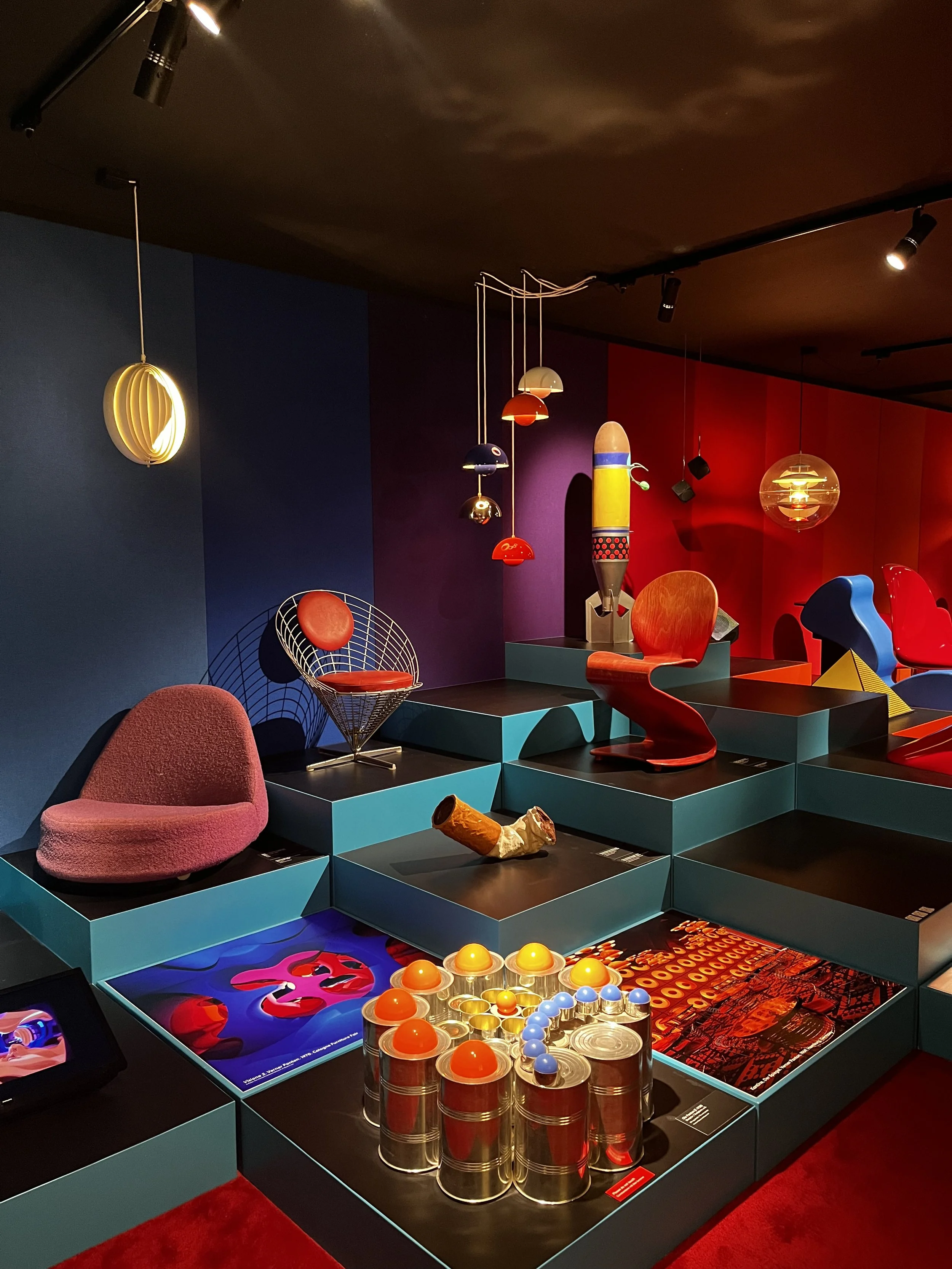 Exhibición de muebles y objetos de diseño moderno en una sala con paredes de colores vivos y suelos negros. Incluye una silla roja, una estructura de mimbre con cojín rojo, y varias lámparas colgantes de diseños innovadores.