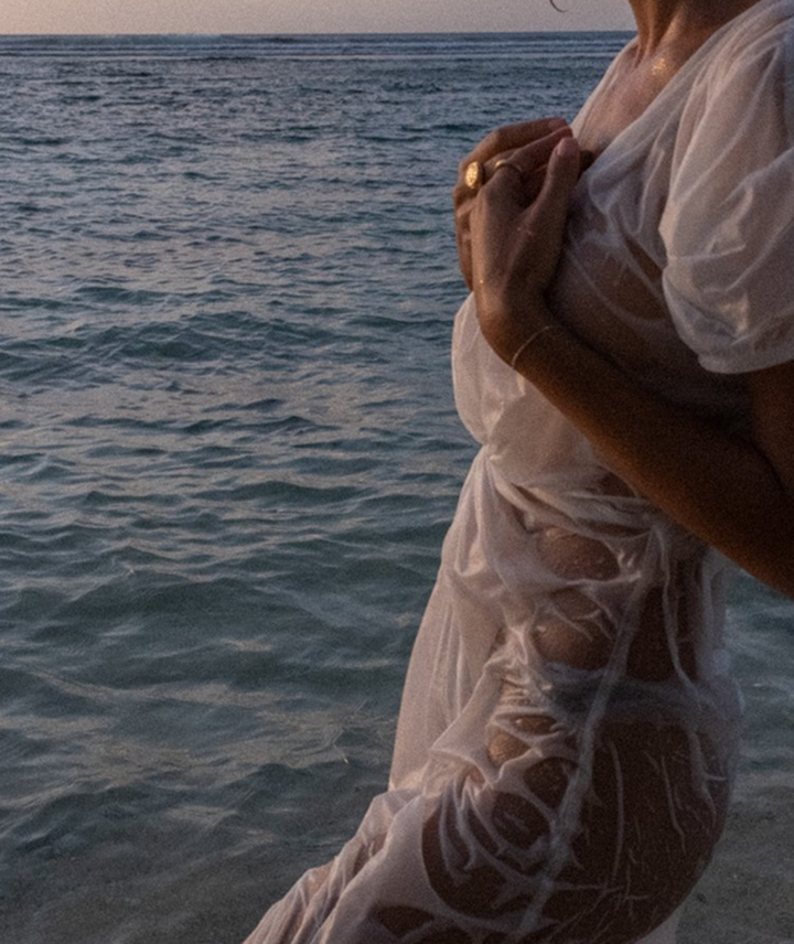 Persona con ropa blanca, parada junto al mar, sosteniendo su pecho con ambas manos.