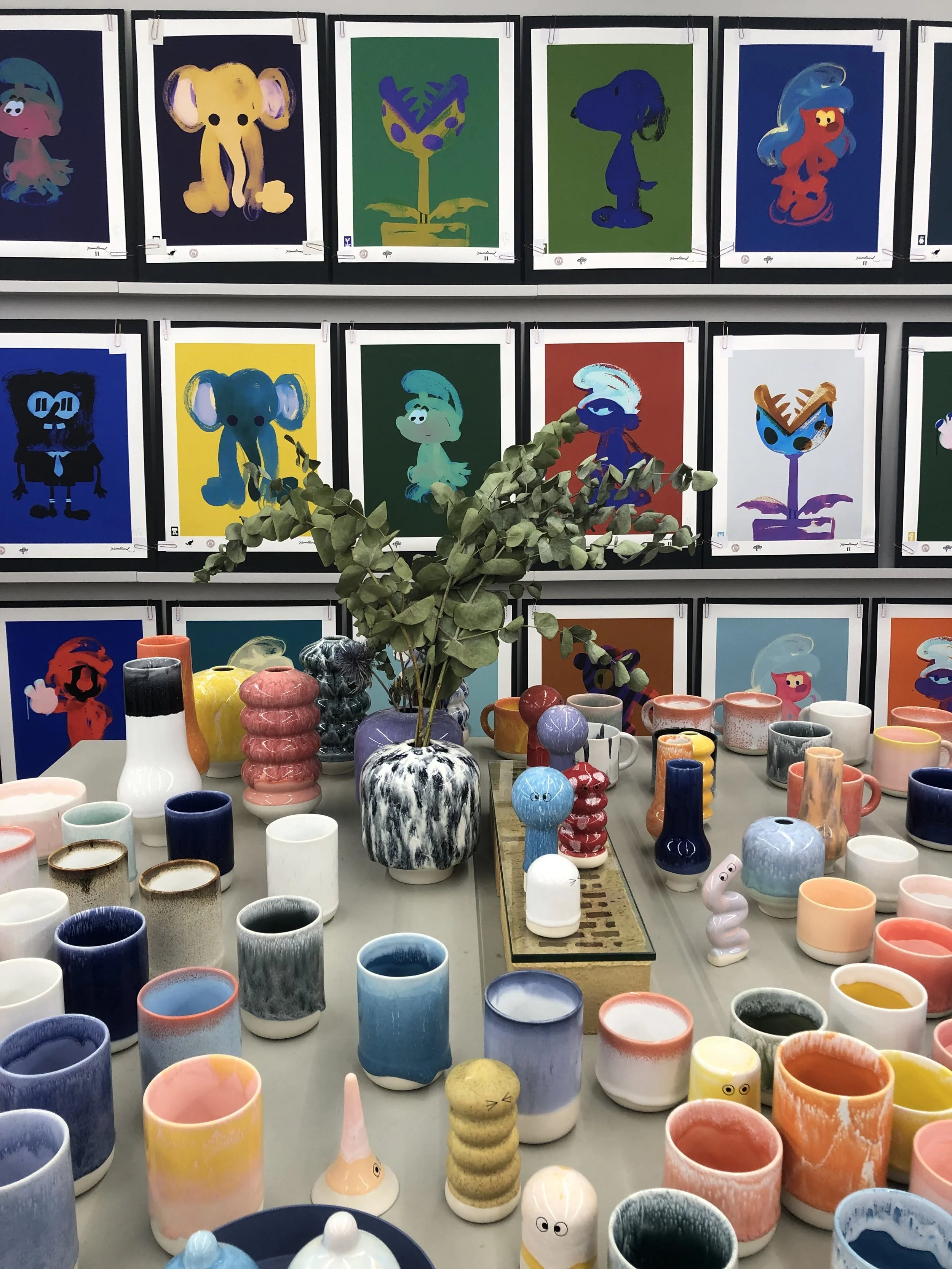 Mesa con varios jarrones y figuras de cerámica, con cuadros de dibujos de personajes de dibujos animados en la pared de fondo.