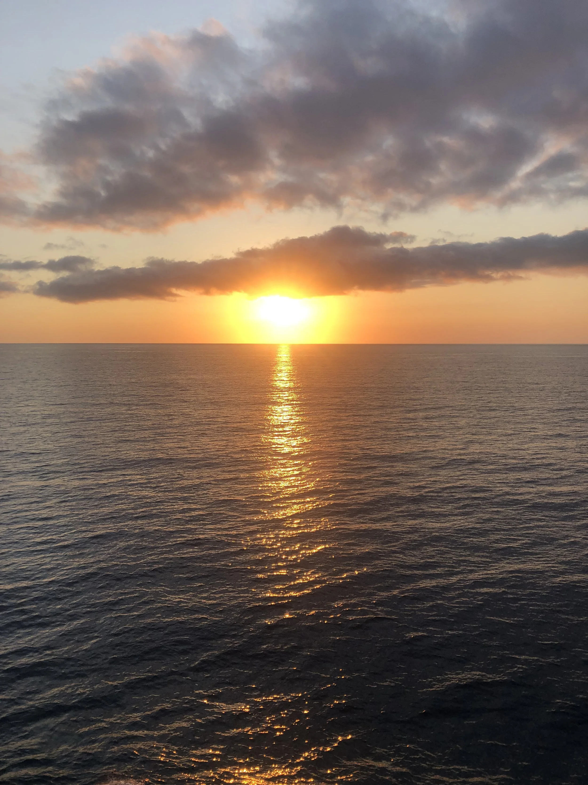 Atardecer sobre el mar, con el sol en el horizonte, reflejándose en las olas y algunas nubes en el cielo.