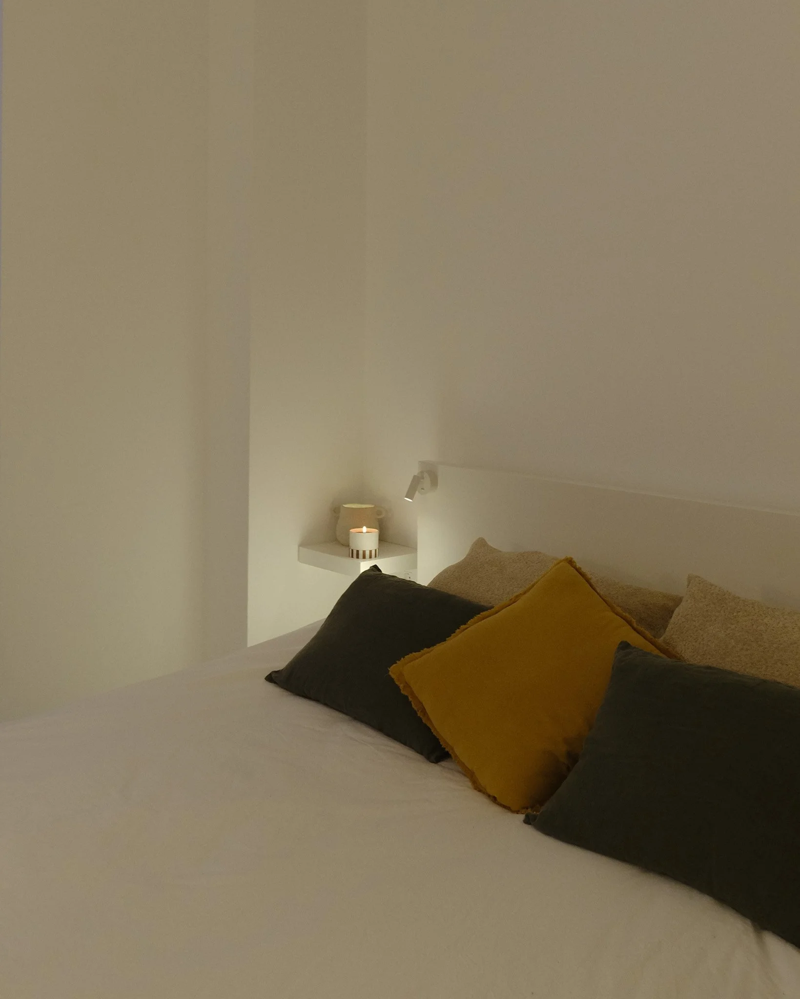 Cama con almohadas beige y negras, velas en estantes blancos, luz de lectura dirigida