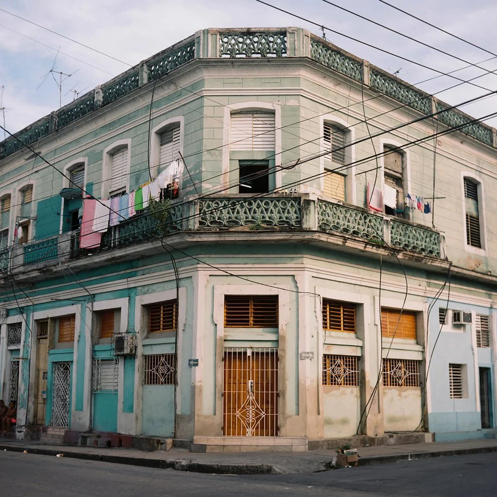 Edificio de tres pisos en una esquina, con paredes verdes y blancas, balcón con baranda de metal y ropa colgada en la cuerda de tendido, cables eléctricos cruzando la calle, y ventanas con rejas y persianas.