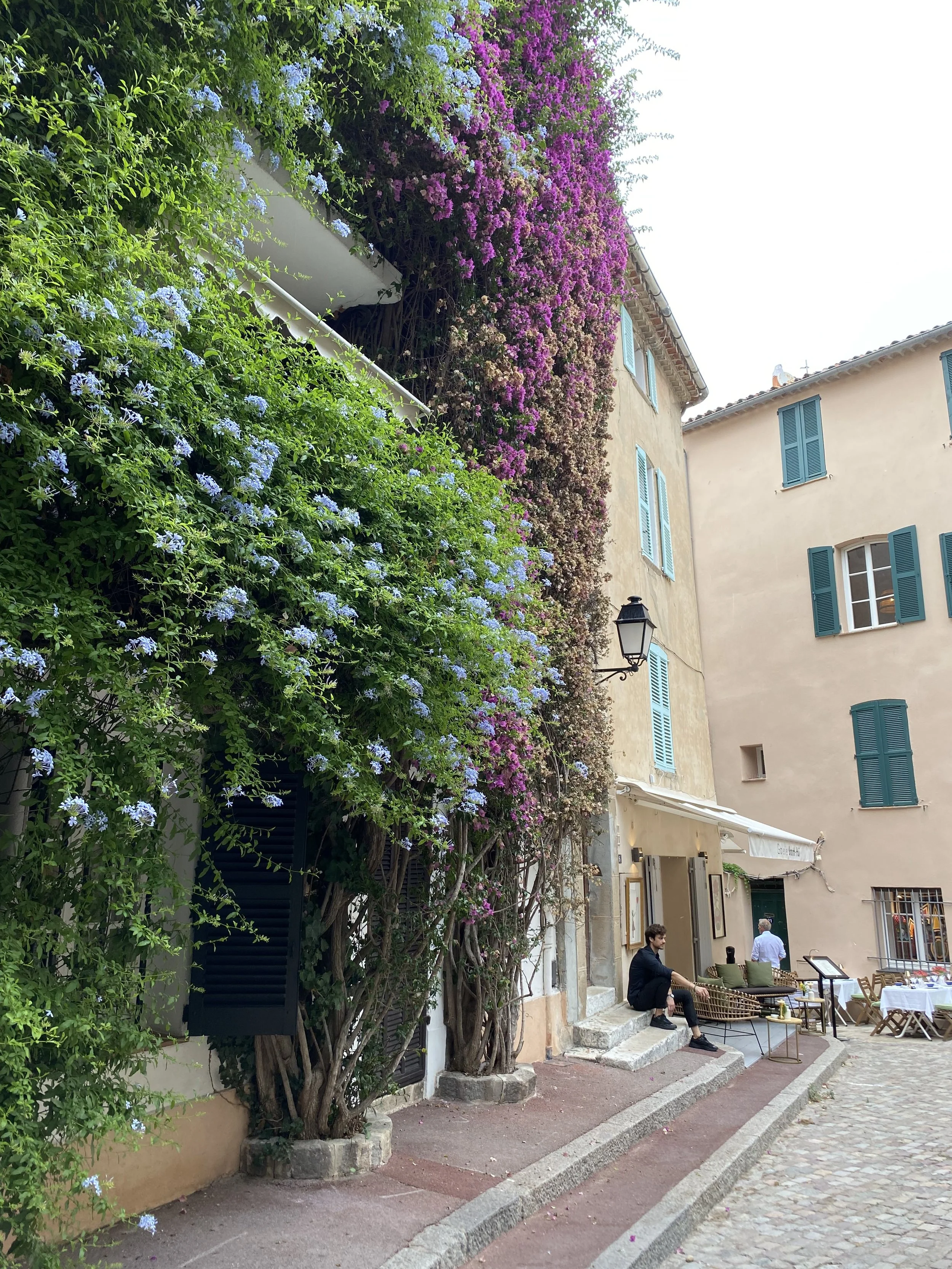 Calles de un pueblo con paredes de casas cubiertas de flores moradas y azules, personas sentadas en la banqueta, y mesas dispuestas para comer afuera, en un ambiente tranquilo y pintoresco.