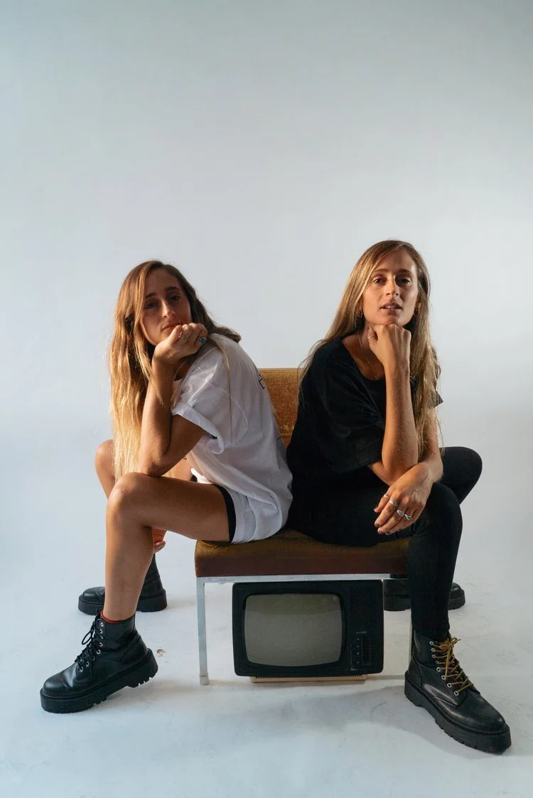 Dos mujeres sentadas en un banco con un televisor viejo debajo, una de ellas lleva blusa blanca y tacones, la otra lleva camiseta negra y botas militares, en un fondo gris.