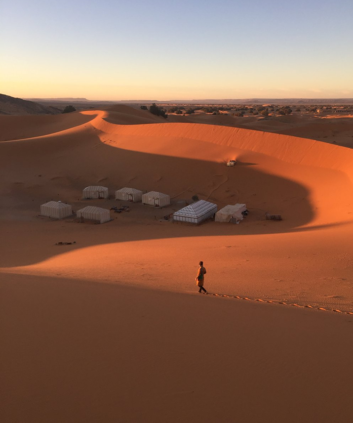 Persona caminando en dunas de arena al atardecer, con varias tiendas y vegetación en el fondo, en un desierto amplio.