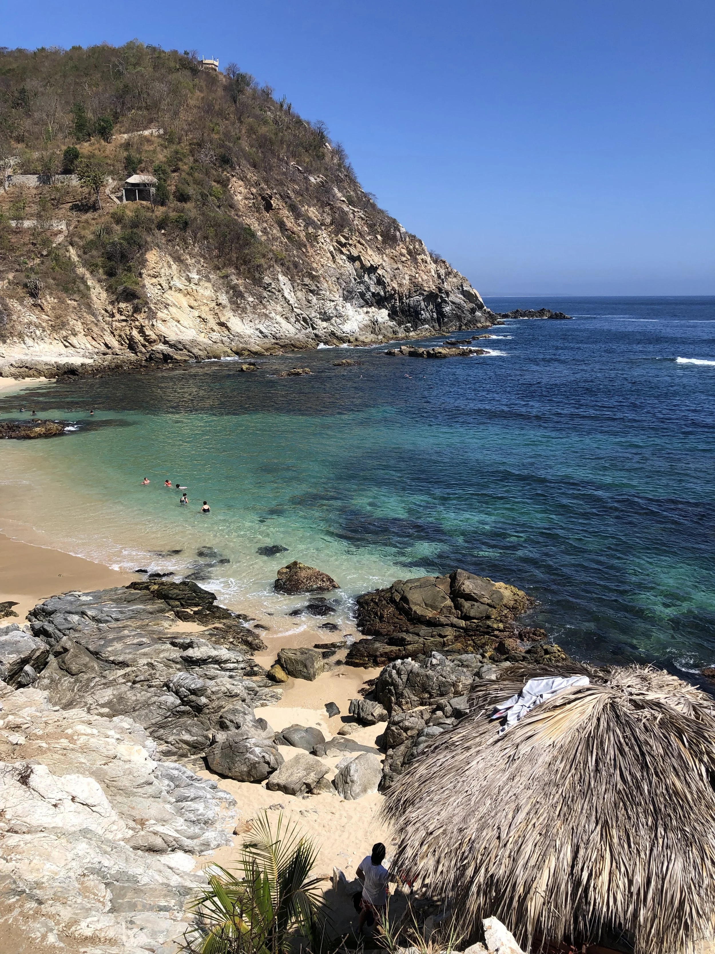 Una playa con agua cristalina en una bahía, rodeada de rocas y un cerro con vegetación y algunas cabañas en la cima, bajo un cielo despejado.