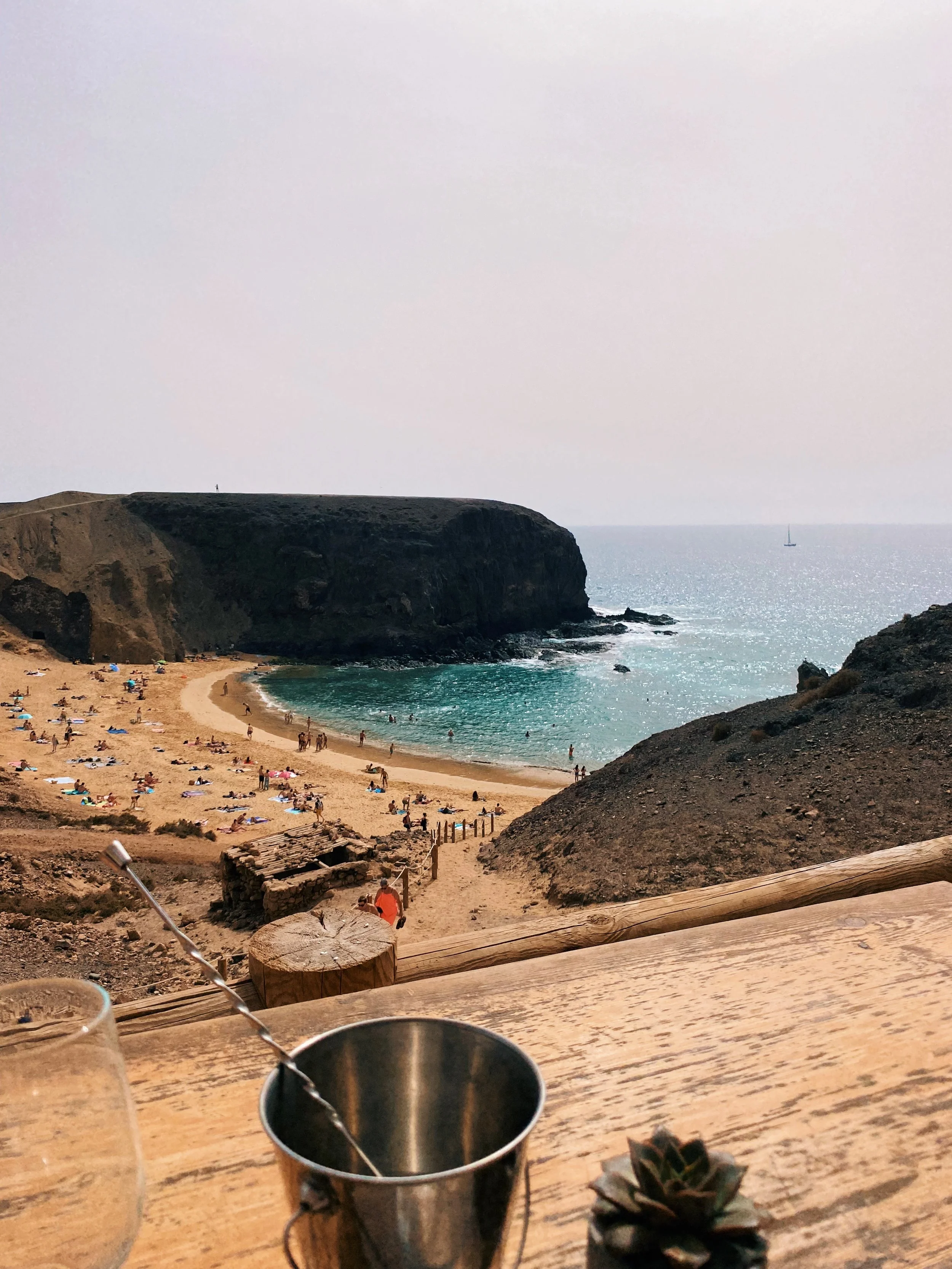 Vista de una playa con arena dorada, rodeada de acantilados rocosos, personas tomando el sol, bañándose en el mar y disfrutando del día, con un pequeño toldo y objetos en primer plano.