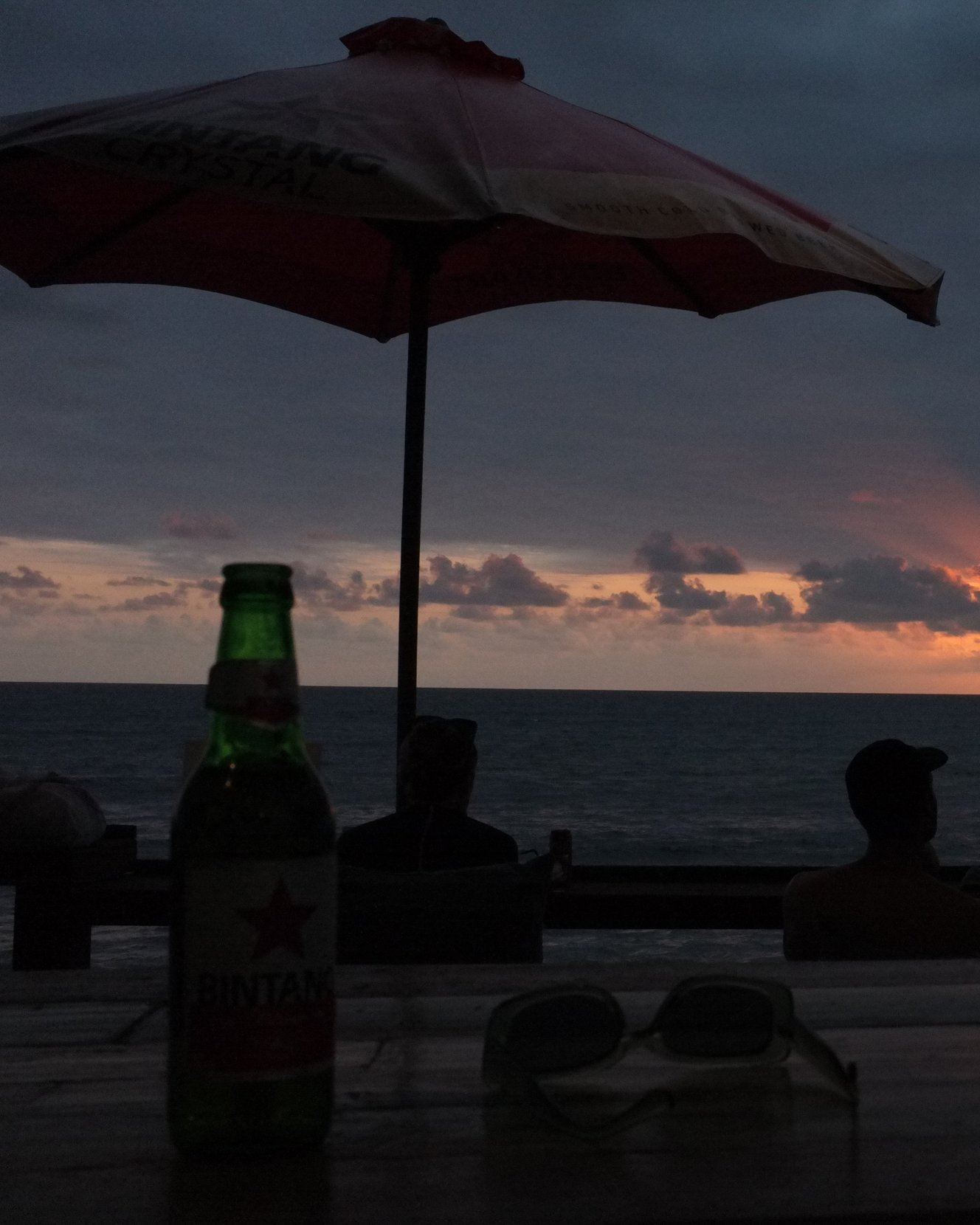 Viendo una puesta de sol en la playa con una sombrilla y una botella de cerveza junto a unas gafas de sol sobre una mesa de madera.