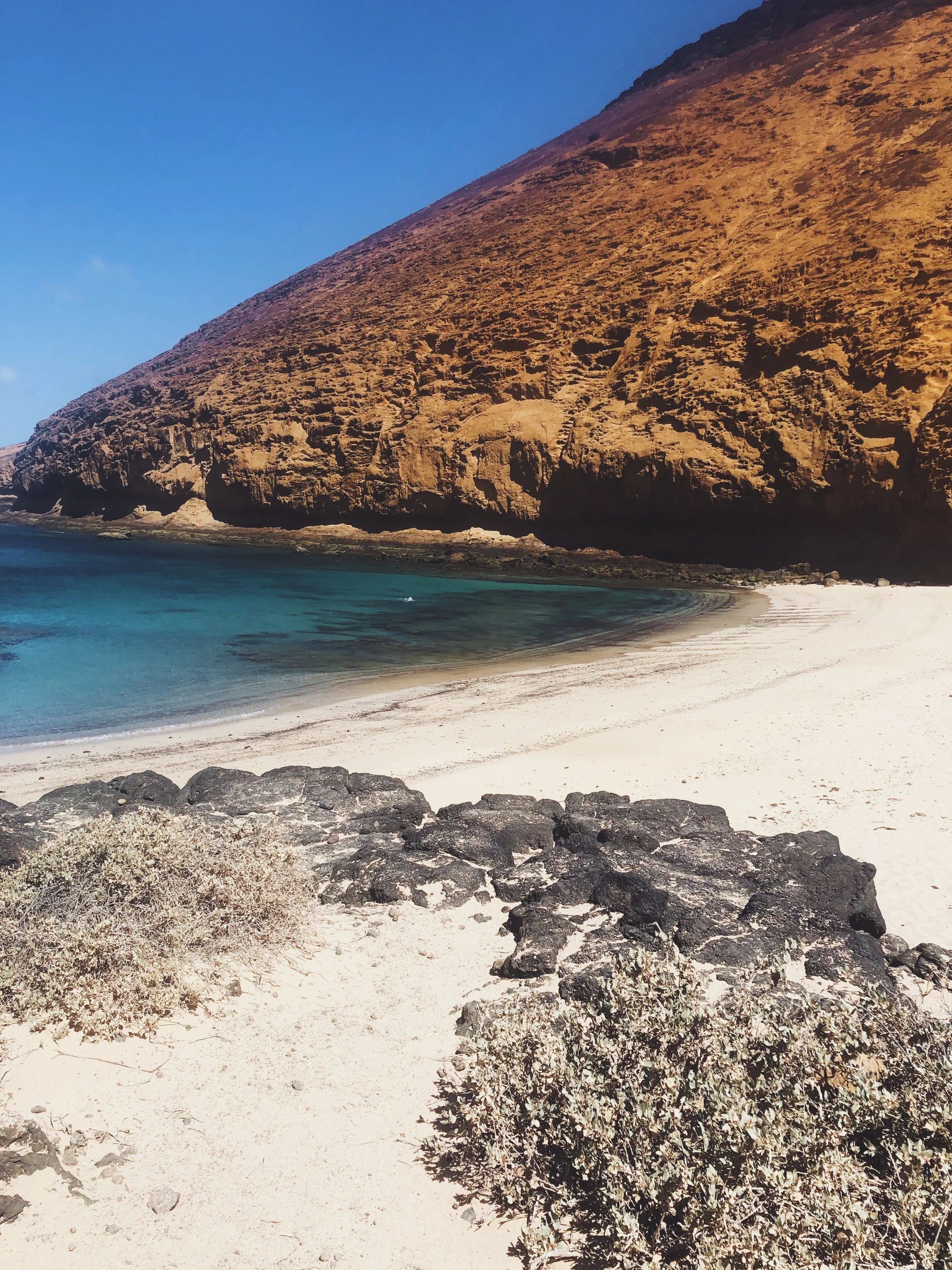 Playa de arena blanca con rocas negras y un acantilado de roca marrón en el fondo, junto al mar azul.