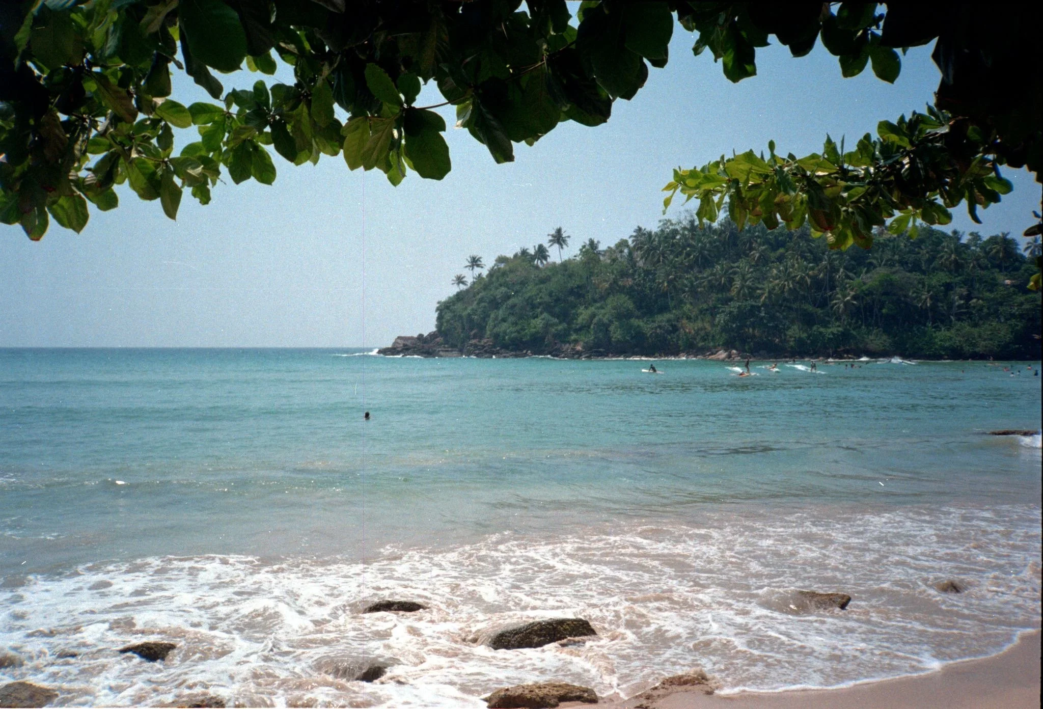 Playa tropical con agua clara, arena y rocas, vista desde la orilla con árboles cubriendo la parte superior, y una colina de palmeras al fondo.