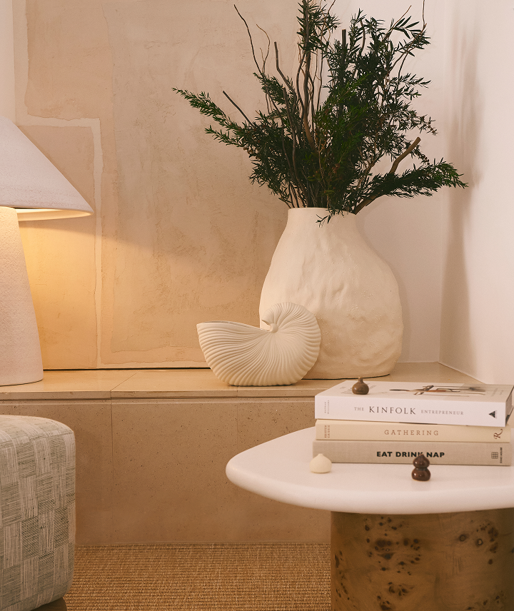 Vasera blanca con plantas verdes y ramitas secas, libros apilados en una mesa blanca, una lámpara y un sillón gris en un ambiente decorativo y acogedor.