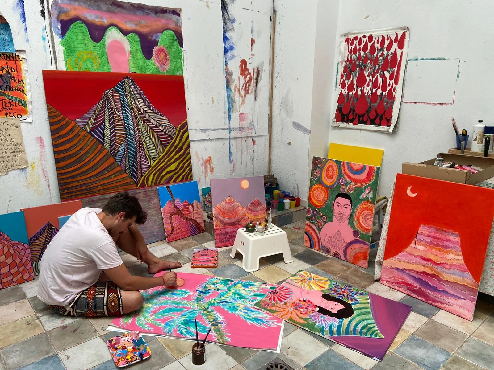 Un hombre sentado en el suelo de un estudio de arte, rodeado de varias pinturas coloridas y en proceso, con paredes blancas y piso de baldosas. Muchas obras de estilo vibrante y abstracto decoran el espacio.