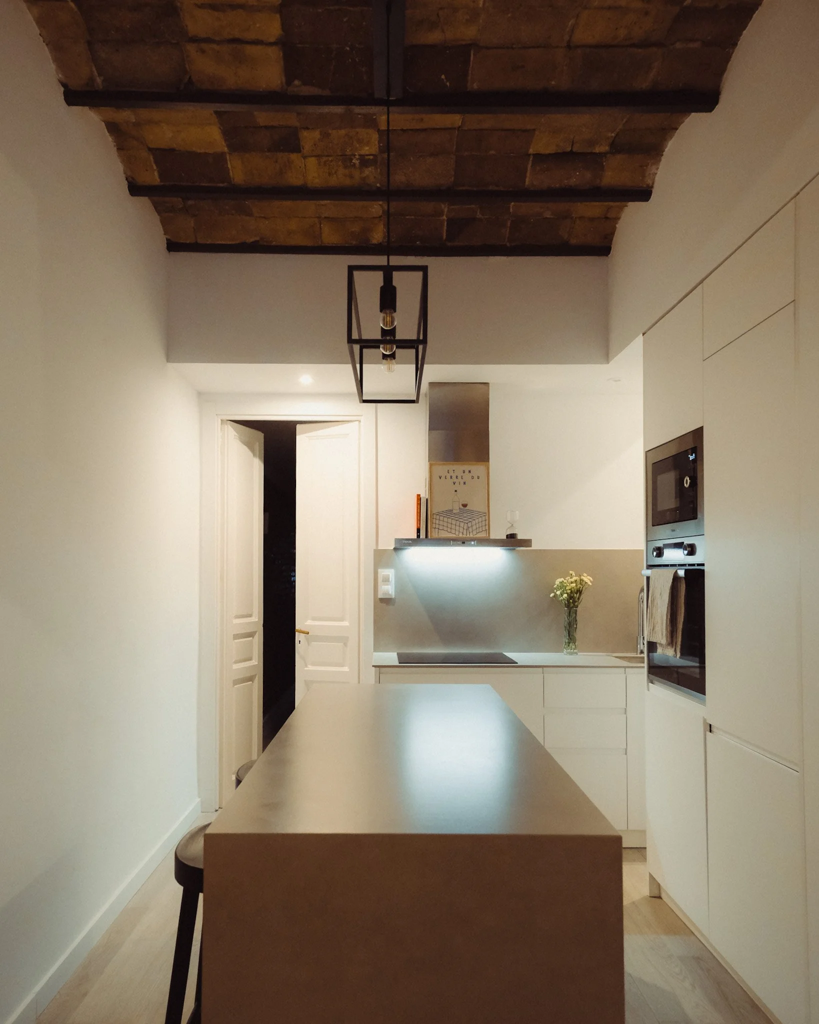 Cocina moderna con techo de ladrillos expuestos, isla de color madera clara, gabinetes blancos, horno empotrado, lámpara colgante de estructura cuadrada, y una lámpara de luz en la pared además de un florero con flores.