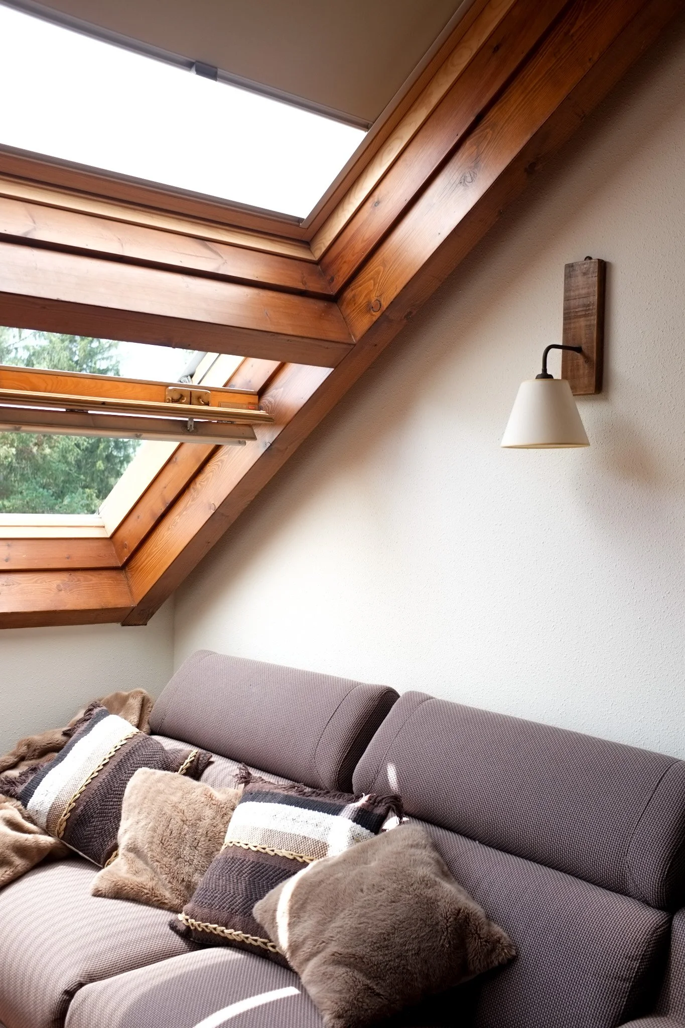 Sala con sofá gris y cojines de diferentes colores y texturas, paredes blancas, ventanas en el techo de madera, y lámpara de pared con base de madera.